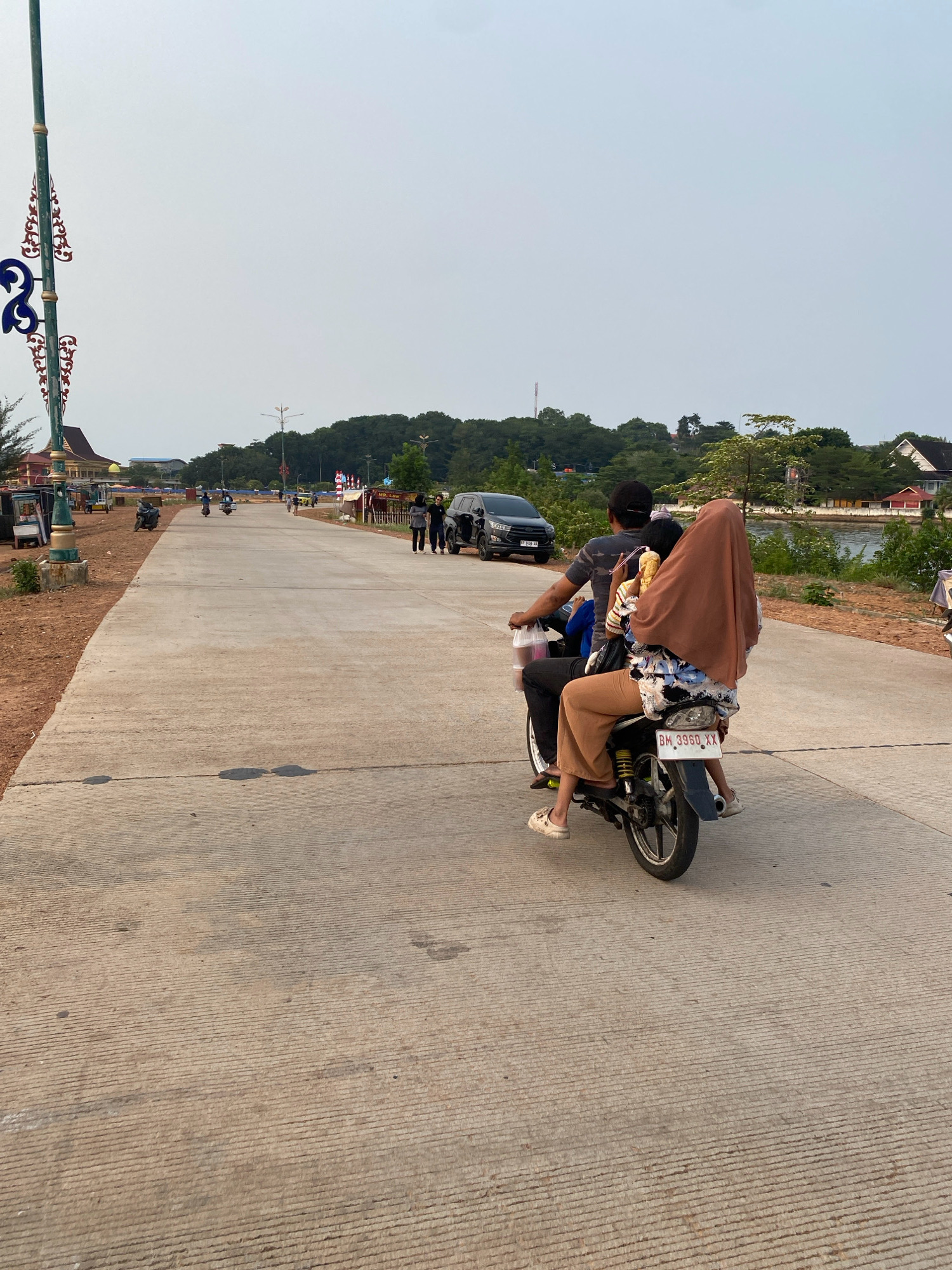 The image depicts a scene on a concrete road in Tanjung Pinang, Indonesia, likely in the vicinity of the Tepi Laut area, given the presence of a body of water and vegetation in the background. The road is made of grooved concrete and appears to be a dual carriageway with a central divide not visible. In the foreground, a man and a woman are riding a motorcycle. The man is driving, wearing a dark t-shirt and a black cap. The woman is seated behind him, covered with a light brown hijab, and appears to be holding a child or a bundle wrapped in fabric. Her legs are clad in light brown pants. The motorcycle has a license plate with the characters "BM 3960 XX".
The environment suggests a somewhat urban or semi-urban setting. On the left side of the road, there are some structures that appear to be stalls or small buildings, and a decorative lamppost with intricate blue and red designs. Further in the background, there are trees, buildings, and a body of water. On the right side of the road, a black SUV is parked, and two people are standing next to it, facing away from the camera. The sky is overcast with a pale, hazy appearance. The lighting suggests it might be late afternoon or early morning. The overall impression is of a typical street scene in a coastal city.