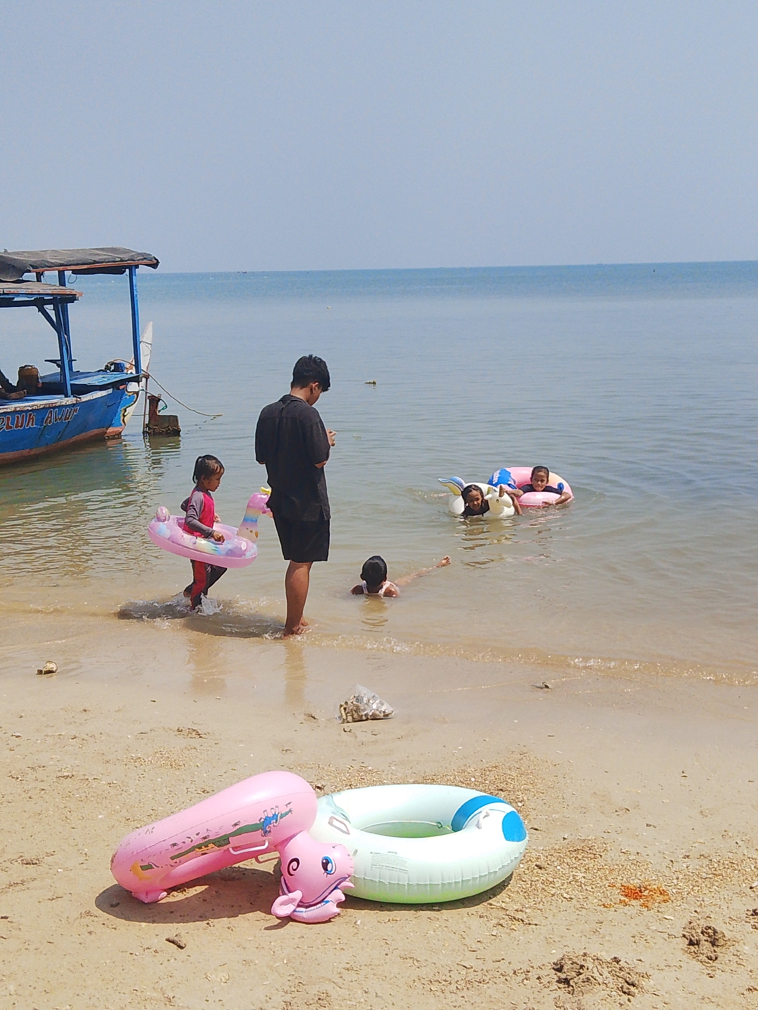 The image depicts a sunny day at the beach in Pejagan, Indonesia. In the foreground, two inflatable pool toys, one pink with a unicorn design and the other a mint green ring with blue accents, lie on the sandy shore. To the left, a blue boat with a covered canopy and the inscription "PLUK AW" is partially visible, anchored in shallow, clear water.
Several people are interacting with the water. A man in a black t-shirt and shorts stands near the water's edge, looking at his phone. A young girl in a pink swimsuit is wading in the water, wearing a pink unicorn-shaped inflatable ring. Further out in the water, two children are playing with inflatable rings; one child is wearing a pink ring, and the other is with a white unicorn-shaped ring. Another child is partially submerged in the water, with their head visible. The sea stretches to the horizon under a clear blue sky. The sand is a light brown, with scattered shells and debris.