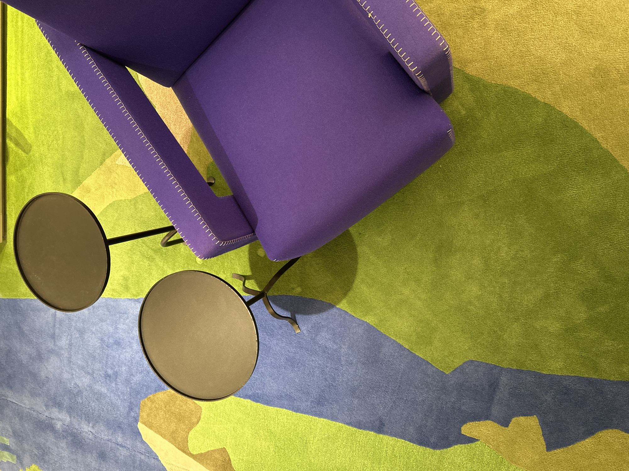 An overhead shot shows a purple chair with visible white stitching along the arms, positioned on a multi-colored carpet featuring shades of green and blue. A small, black table with two circular surfaces stands to the left of the chair. The exact location within London, United Kingdom is not discernible from the image. No people are visible.