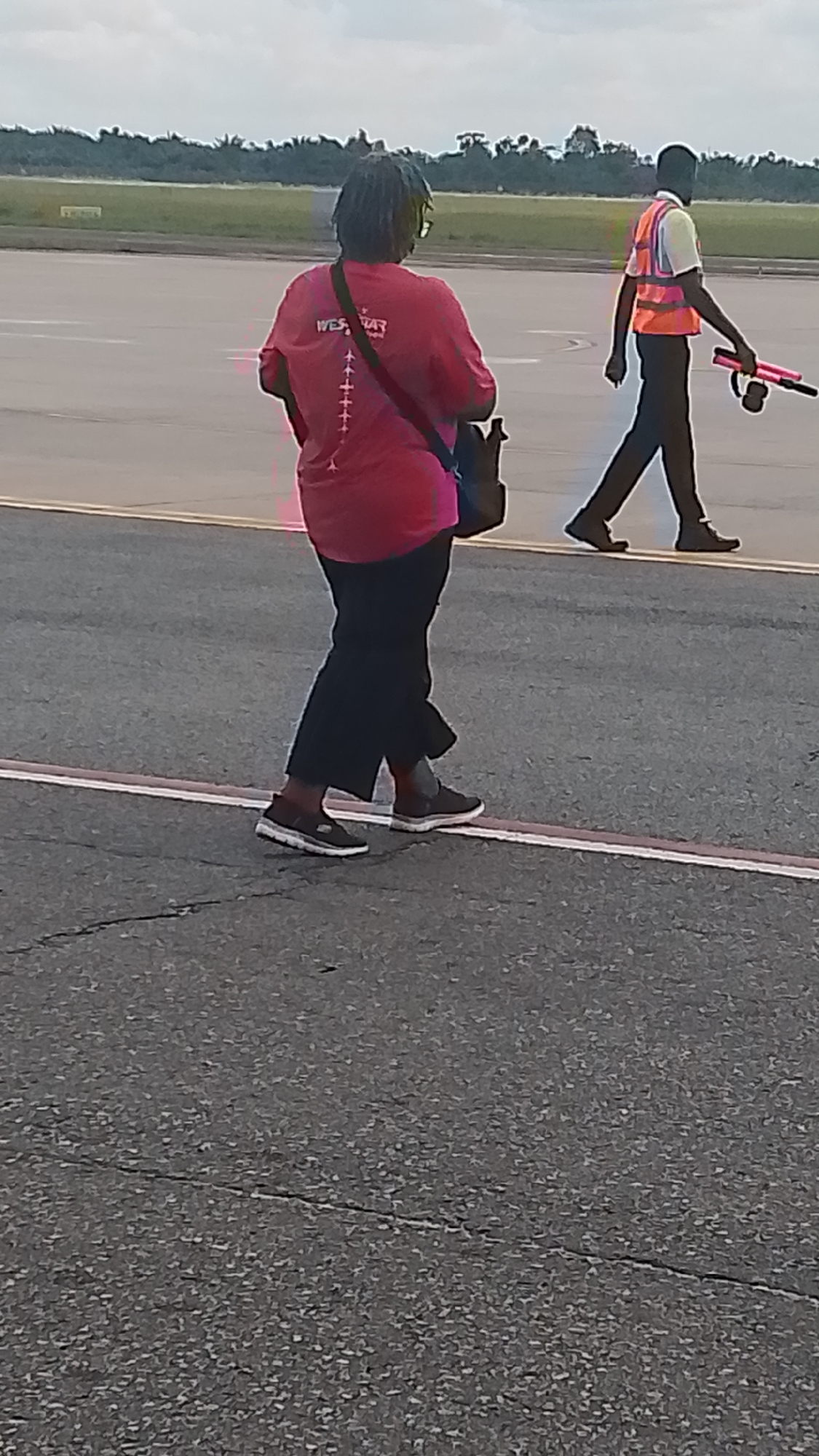 The image shows two individuals walking on an airport tarmac. The person in the foreground is wearing a bright pink t-shirt with a graphic design on the back that includes the text "WEST AIR" and several small airplane icons. They are also wearing black pants and dark sneakers. A dark bag is slung over their shoulder. The second person, further in the background, is wearing a high-visibility orange vest over a light-colored shirt and dark pants. They are carrying a pink object with a black handle, possibly a signaling device or tool. The background features an open expanse of tarmac, with a line of trees visible in the distance under a cloudy sky. The scene appears to be outdoors during the daytime, likely at an airport given the setting and the presence of airport personnel. The overall mood is neutral, depicting everyday activity at an airport.