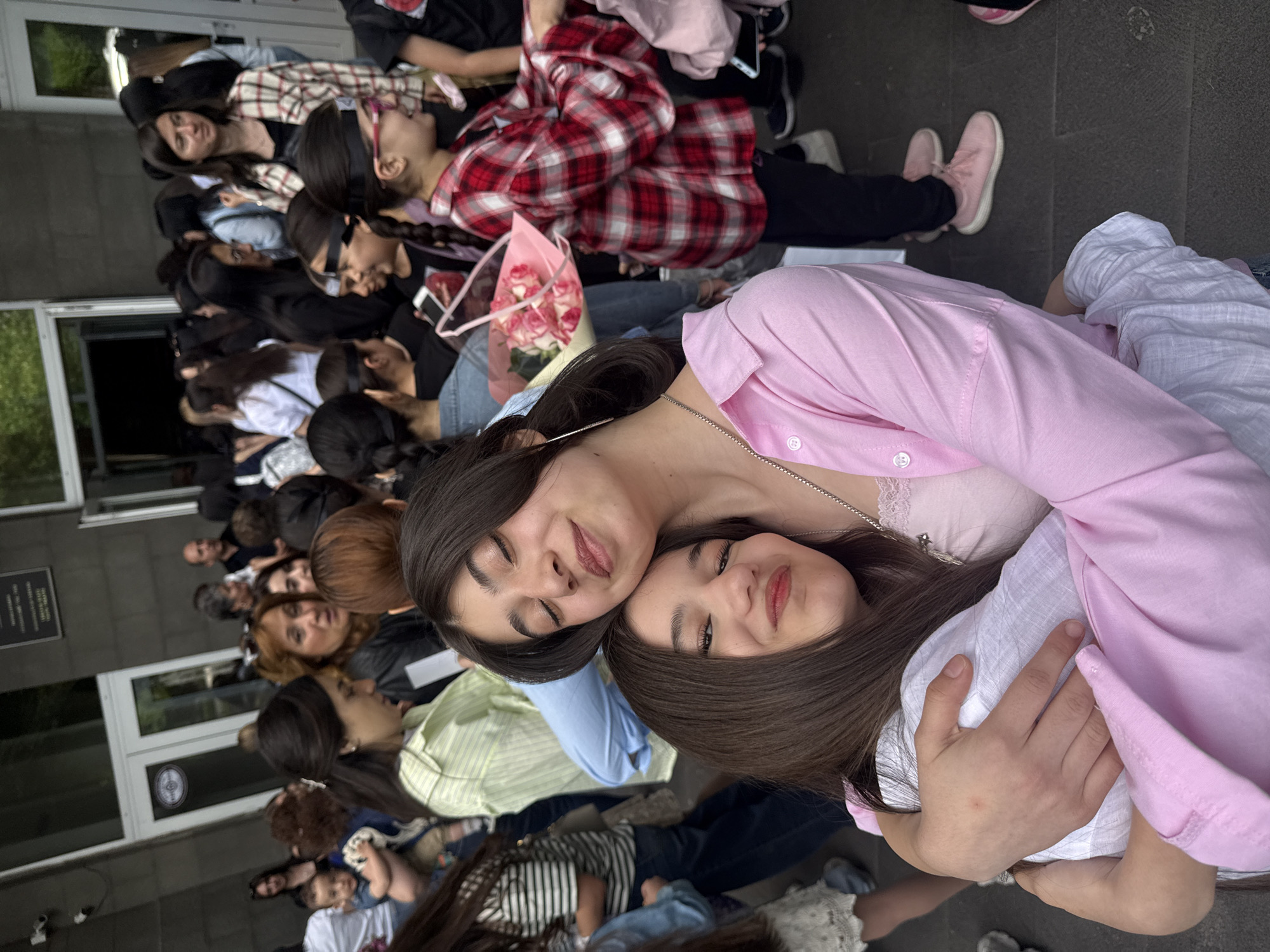A crowded outdoor scene in Yerevan, Armenia, captures a group of people, predominantly young women, sitting and posing for a photo. Two women in the foreground are embracing, one in a pink shirt and the other in a white shirt. Behind them, numerous individuals are seated on what appears to be pavement, facing in the same direction. Some are wearing masks or face coverings. An assortment of casual clothing styles is visible, including plaid shirts, jeans, and dresses. A bouquet of pink flowers is among the group. In the background, a building with white doors and a dark sign is visible. The overall impression is that of a group gathered for an event, possibly a graduation or celebration.