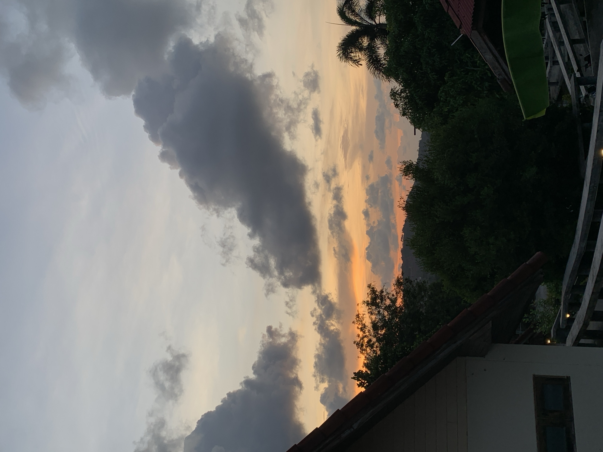 The image captures a sunset scene in บ้านบางรักษ์, Thailand. Dark, heavy storm clouds dominate the sky's left side, contrasted by a vibrant orange and yellow sunset in the middle and right portions of the sky. Silhouetted trees, including a palm tree, and parts of buildings frame the sunset. The buildings have red tile roofs, and there are wooden structures and green foliage nearby. The scene is shot from a low angle, looking up towards the sky. The overall impression is of a dramatic and beautiful sunset amid a tropical landscape.