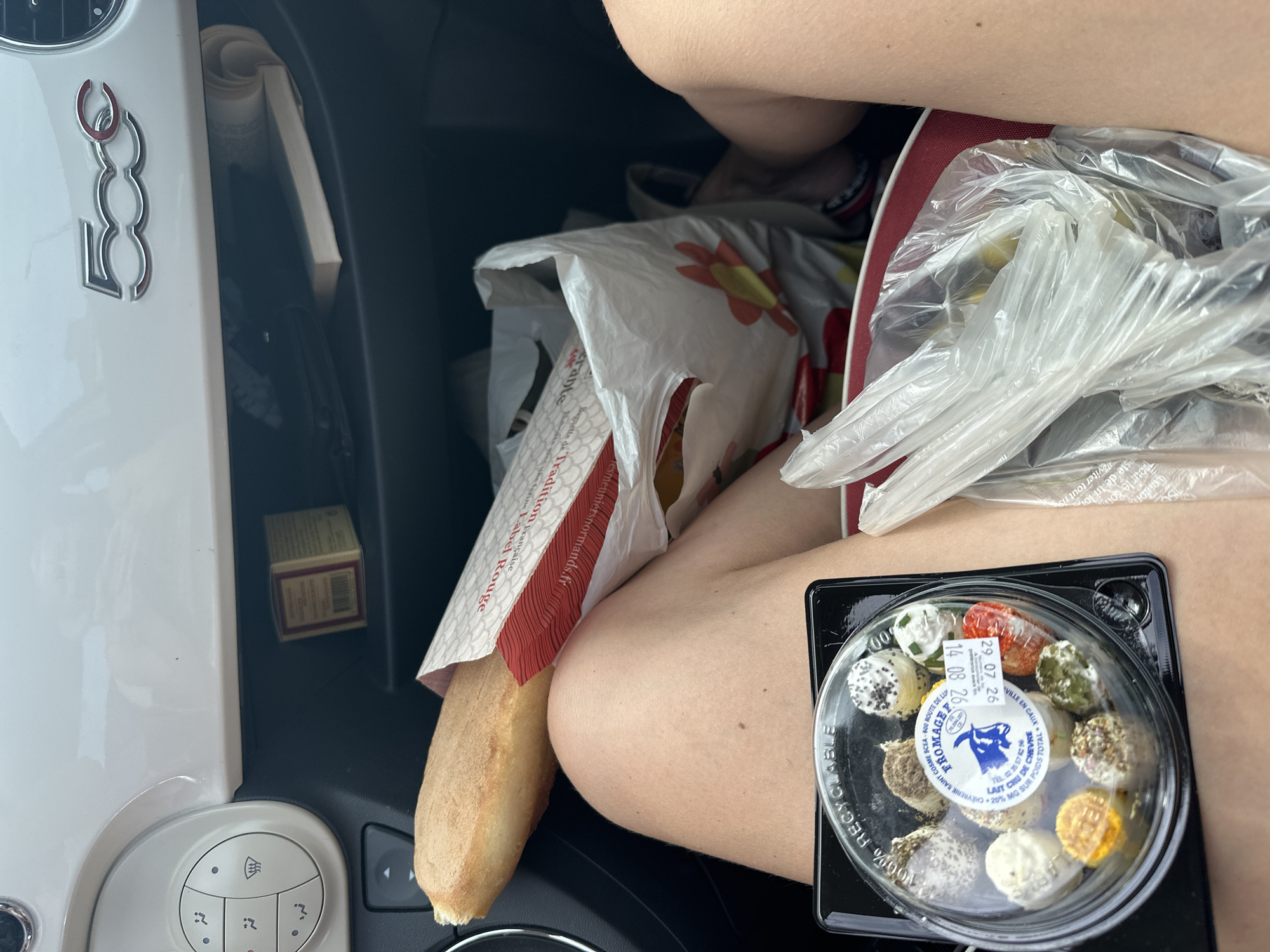 The image is a close-up, eye-level shot taken inside a Fiat 500 car, likely in Ouville-la-Rivière, France, given the text on the baguette packaging. In the foreground, a person's bare legs are visible, positioned as if sitting in the driver's or passenger's seat. A black plastic container filled with small, assorted cheeses is placed on the person's lap. Next to the cheese, a clear plastic bag contains what appear to be green olives. Behind the person, a white plastic shopping bag with a red and yellow flower design holds a baguette, which protrudes from a red and white paper wrapper. The car's interior is visible, including the dashboard with the "500" logo, climate control buttons, and a glove compartment containing a small box and a rolled-up document. The scene suggests a casual stop, possibly for a picnic or a snack, with groceries purchased from a local market or boulangerie.