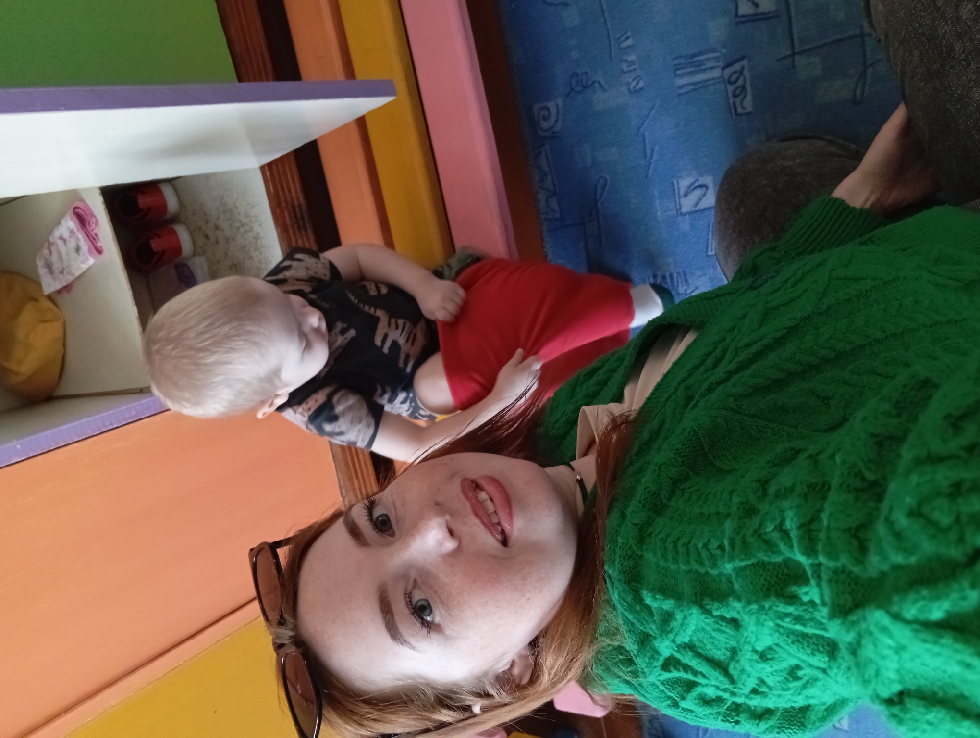 A selfie shows a woman with red hair wearing a green sweater and sunglasses, lying on the floor with a toddler wearing a black shirt and red pants. The toddler appears to be reaching into a shelf containing red shoes and other items. The floor is carpeted with a blue patterned rug. A portion of the woman's leg and denim pants are visible. The location is in Unknown, Unknown.