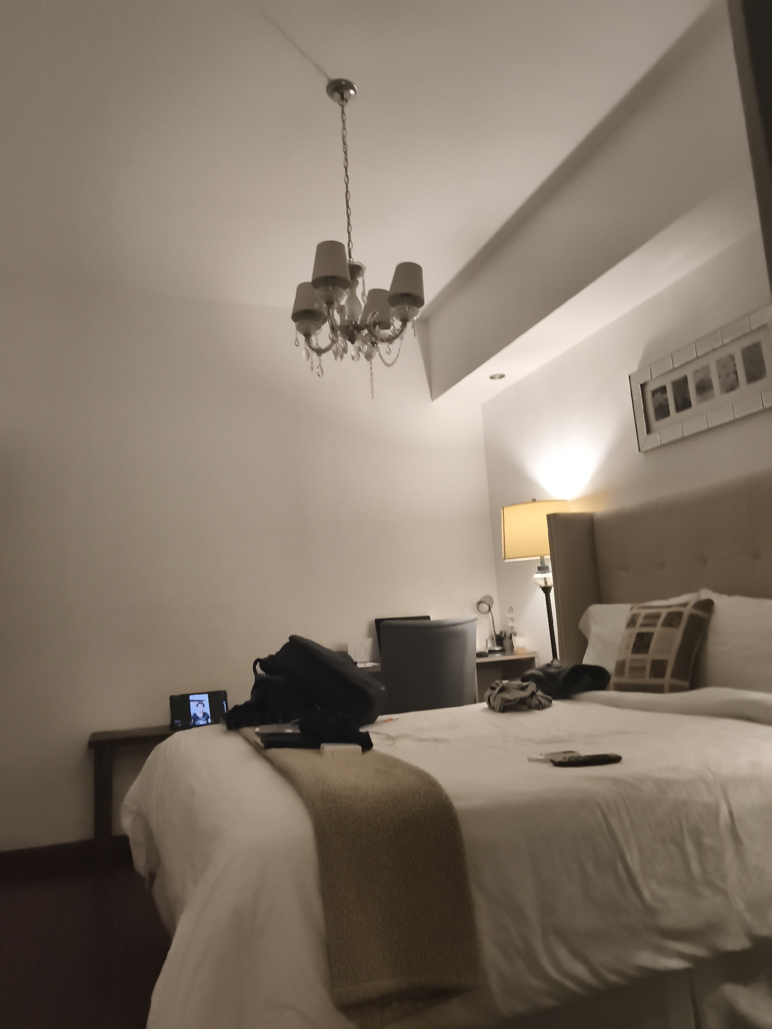 The image shows a bedroom interior. A bed dominates the scene, covered in white linens and topped with a light brown blanket. Pillows are present on the bed, including one with a patterned design. Various personal items are scattered on the bed, such as a black backpack and a remote control. A smartphone stands on a nightstand to the left of the bed. A desk and chair are visible in the background, along with a lamp. The room is lit by a chandelier and the bedside lamp. A picture frame hangs on the wall above the bed's headboard.
While it isn't possible to pinpoint the location as specifically Guatemala City, Guatemala, from the given image, the provided information indicates the photograph was taken there.