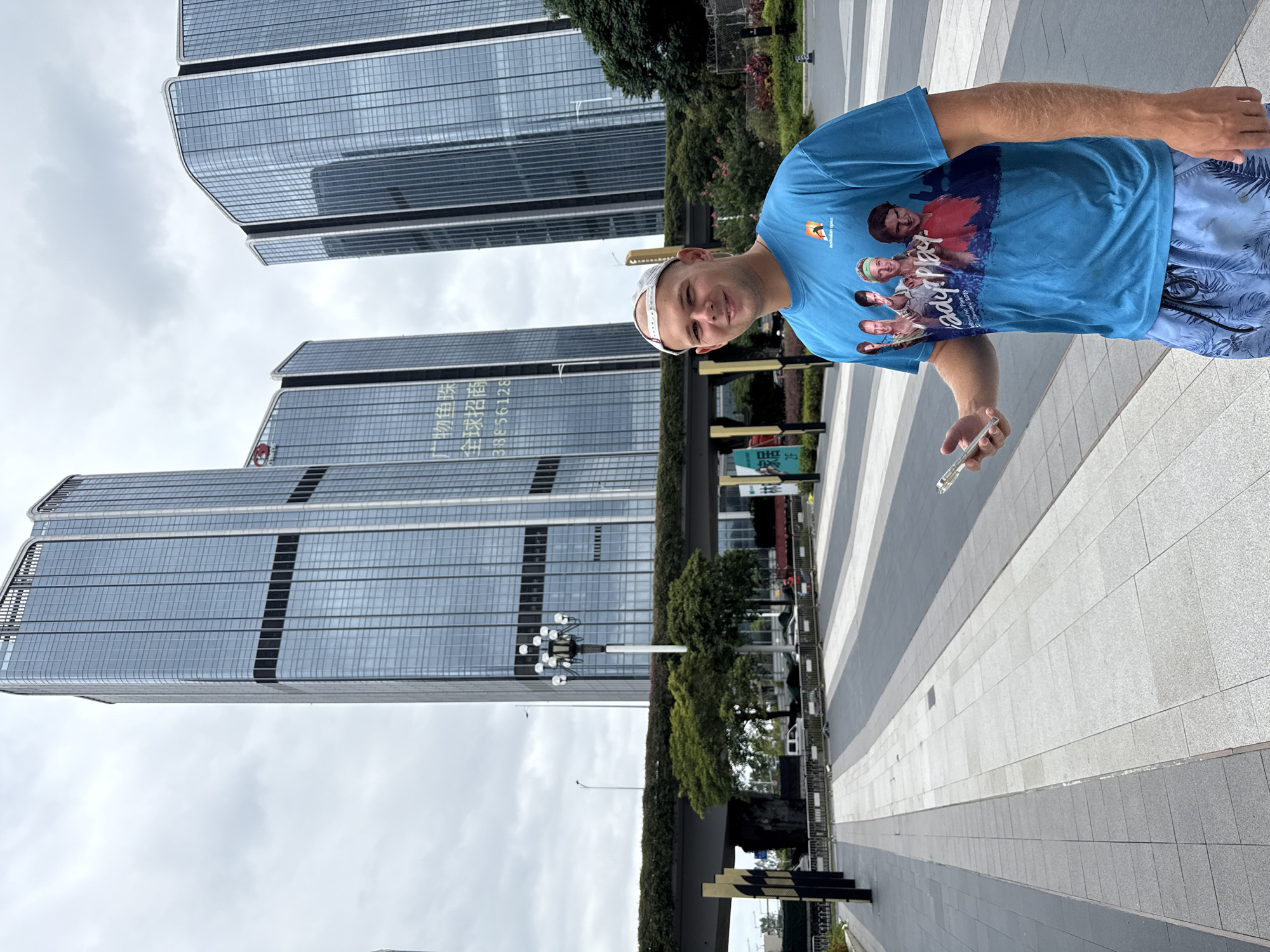 A man in a blue t-shirt and patterned shorts stands on a paved walkway in front of several modern, glass-facade skyscrapers. He holds a smartphone in his right hand. The t-shirt features a graphic of several people and the text "Ready Play Australian Open 27 January 2019." In the background, a multi-level bridge covered in greenery spans across the scene, with trees visible beneath it. The sky is overcast. The architecture suggests a contemporary urban environment, consistent with modern developments in the Huangpu District of China.
