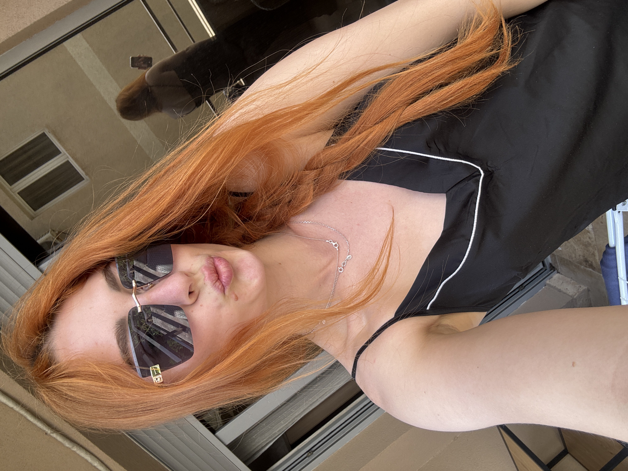 A woman with long red hair, wearing sunglasses and a black top with white trim, is taking a selfie. She is outdoors, possibly on a balcony or patio. The reflection in her sunglasses shows a building and greenery. The specific location in Budva, Montenegro is not determinable from the image.