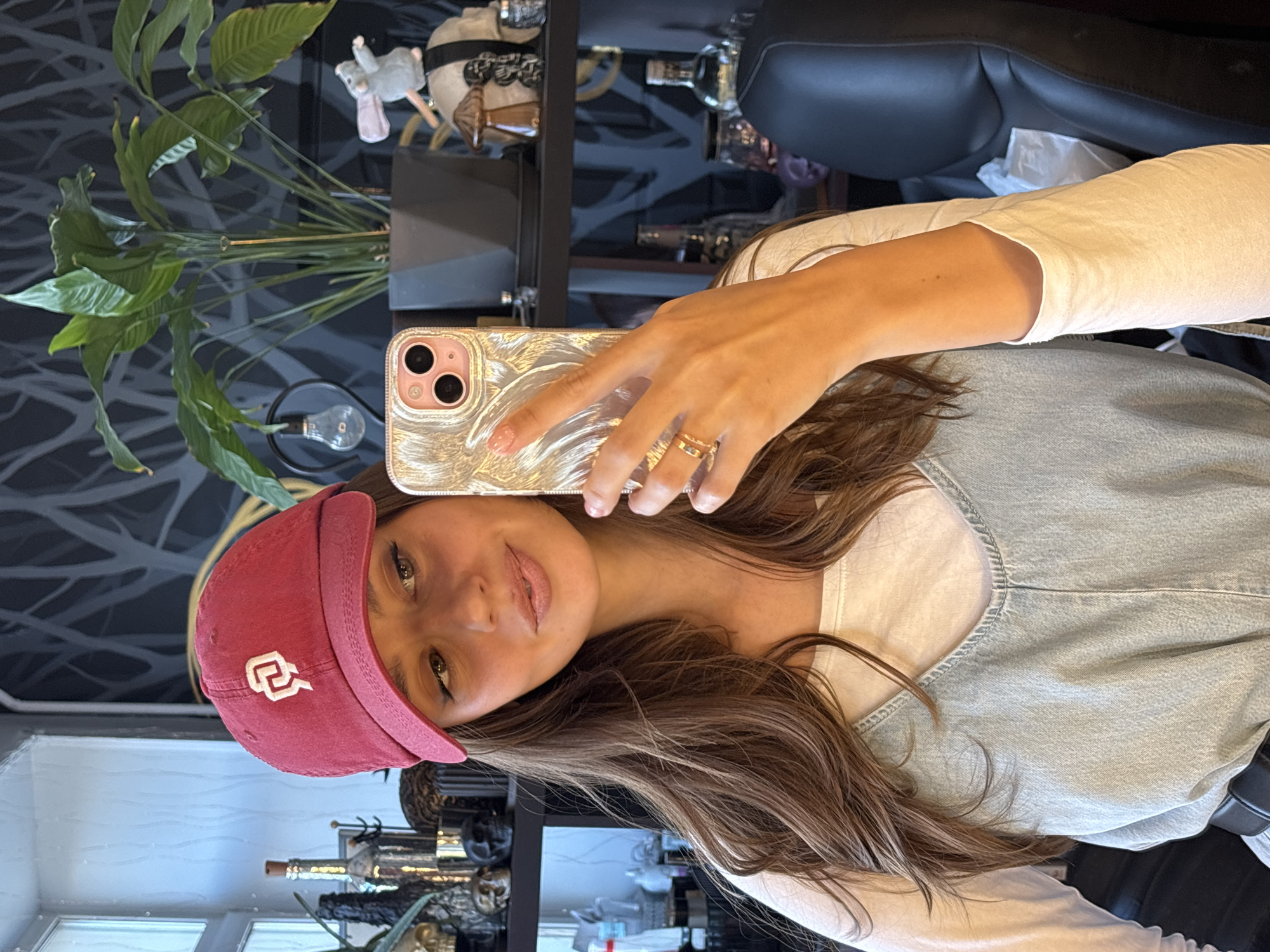 A young woman wearing a red baseball cap with a white emblem, a white long-sleeved shirt, and a light blue denim vest is taking a selfie. She is holding a gold iPhone in her right hand. Her hair is long and brown.
Behind her, there's a shelf with various decorative objects and bottles, along with a green plant. There is also a black leather chair visible. A window with a view of a city is in the background, although the city cannot be identified.
Given the lack of identifying landmarks in the provided image, determining the exact location in Krasnoyarsk, Russia is impossible.