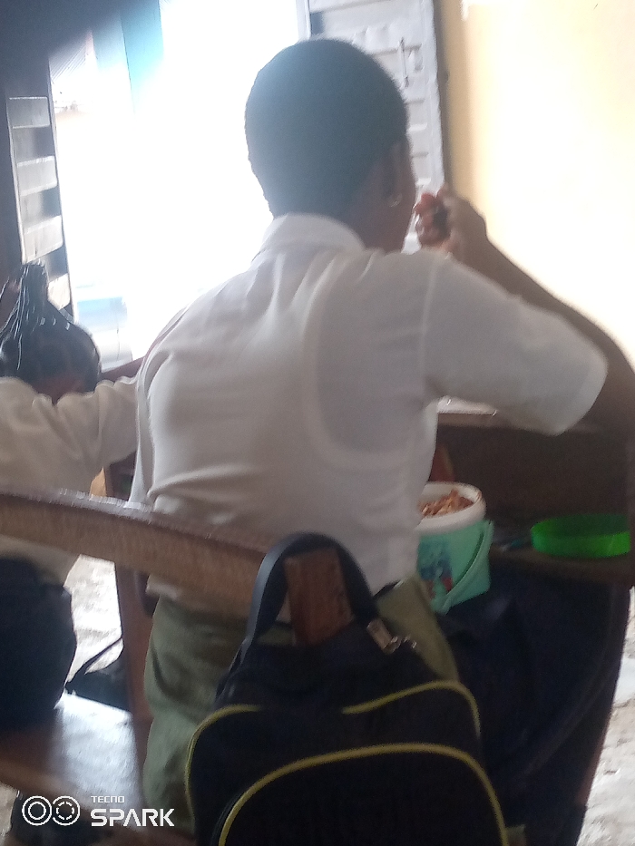 The scene, located in Ajaokuta, Nigeria, depicts an indoor setting with two individuals. A primary subject is viewed from the back, wearing a white short-sleeved shirt and light-colored trousers, seated on what appears to be a wooden bench. Their right hand is raised towards their mouth, indicative of eating from a light blue or green bucket with a red lid, containing light-colored granular food, positioned to their right. A black backpack with yellow trim rests in the lower foreground. To the left and slightly behind the primary subject, a second individual is partially visible, wearing a white shirt and possessing dark, braided hair. The background features a bright, overexposed window with horizontal slats on the left side, serving as the main light source. A horizontal wooden surface extends across the mid-ground, and a separate green circular lid is visible on a surface to the far right.