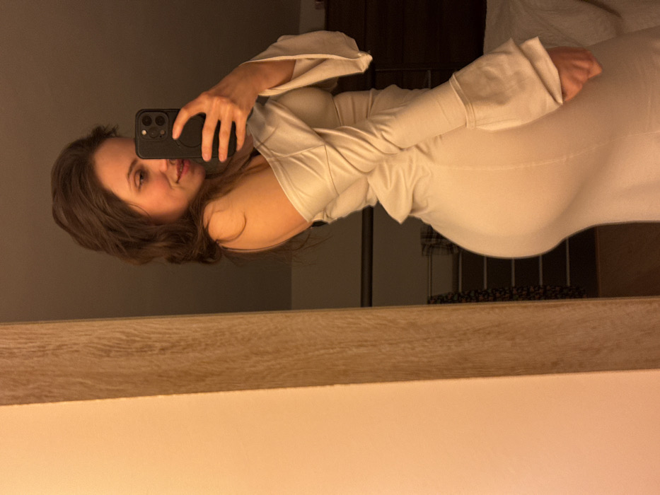 The image displays a mirror selfie of a female individual located indoors in Cava de' Tirreni, Italy. The subject, a woman with long, dark, wavy hair, is depicted from the waist up, facing partially left towards her reflection. She is wearing a light-colored, form-fitting, off-the-shoulder dress with long, bell-style sleeves; a dark strap is visible on her left shoulder. Her right hand holds a dark smartphone, featuring a triple camera array, which is positioned to capture the reflection. She exhibits a subtle smile and directs her gaze towards the phone's lens in the mirror. The background, visible in the mirror's reflection, includes dark wooden furniture, a segment of a white bedspread or pillow, and a metallic towel rack with a dark, patterned cloth draped over it. The lighting within the environment is dim and warm, suggesting an interior setting, likely a private room.