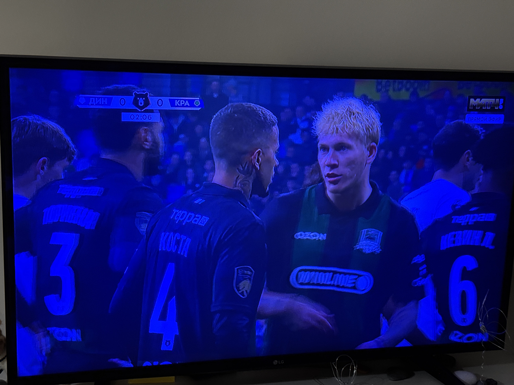 The image shows a television screen displaying a soccer game. The score is tied 0-0 at the 2:06 mark. The game appears to be between teams "Дин" and "КРА". Players wearing jerseys with the numbers 3, 4, and 6 are visible. "KOCTA" is printed on the jersey with number 4. The television has an LG logo. The broadcast has a "MATH!" logo and is labeled "Прямой эфир" (live broadcast). On the jersey with number 6 are the words "терра" and "ИВАНЯХИИ" and "OZON" is visible on both jerseys with numbers 4 and 6. The city and country, Izhevsk, Russia, are not visible within this image.