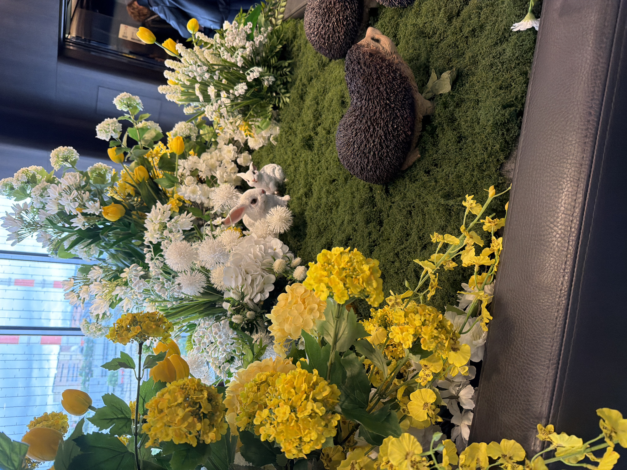 The image shows a floral arrangement, likely indoors, featuring a variety of yellow and white flowers. Among the flowers are two small white rabbit figurines and two hedgehog figurines on a base of green moss. The arrangement is positioned adjacent to a dark gray leather object, possibly a piece of furniture. The scene appears to be taken near a window, through which a cityscape is visible. The location is identified as Moscow, Russia, based on the visible architecture.