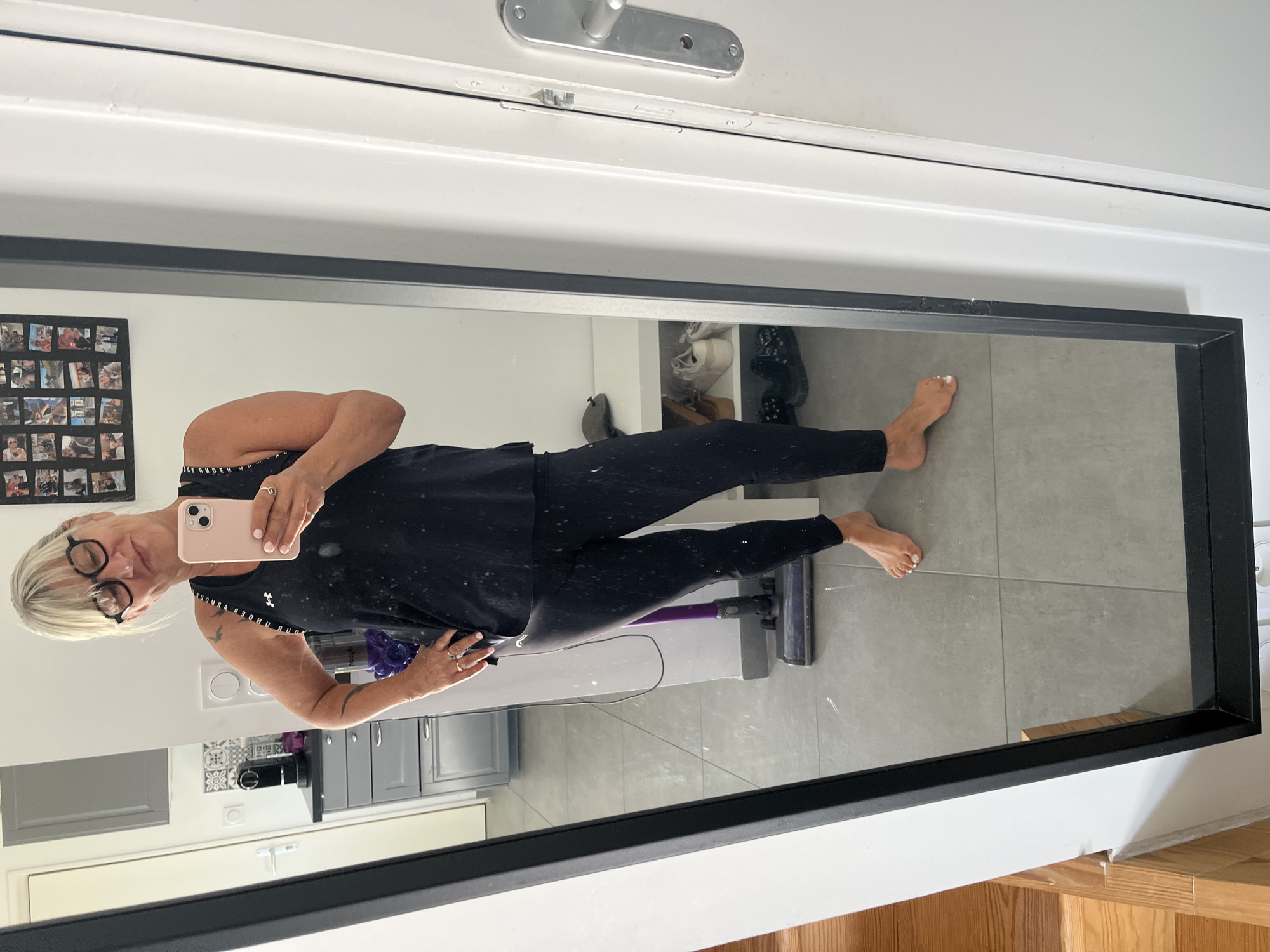 A woman stands in a domestic setting, reflected in a full-length mirror. She is wearing a black tank top and black leggings. She holds a pink smartphone in her right hand, taking a selfie. Her left hand is on her hip. She has blonde hair and wears glasses. Behind her, visible in the reflection, is a kitchen area with cabinets and a coffee machine. To the right of the mirror is a white door with a silver handle. The floor is tiled. There are several pairs of shoes on shelves in the background, and a purple vacuum cleaner is visible on the floor. The image appears to have been taken in an interior space, likely a home, within Chambœuf, France, based on the provided context.