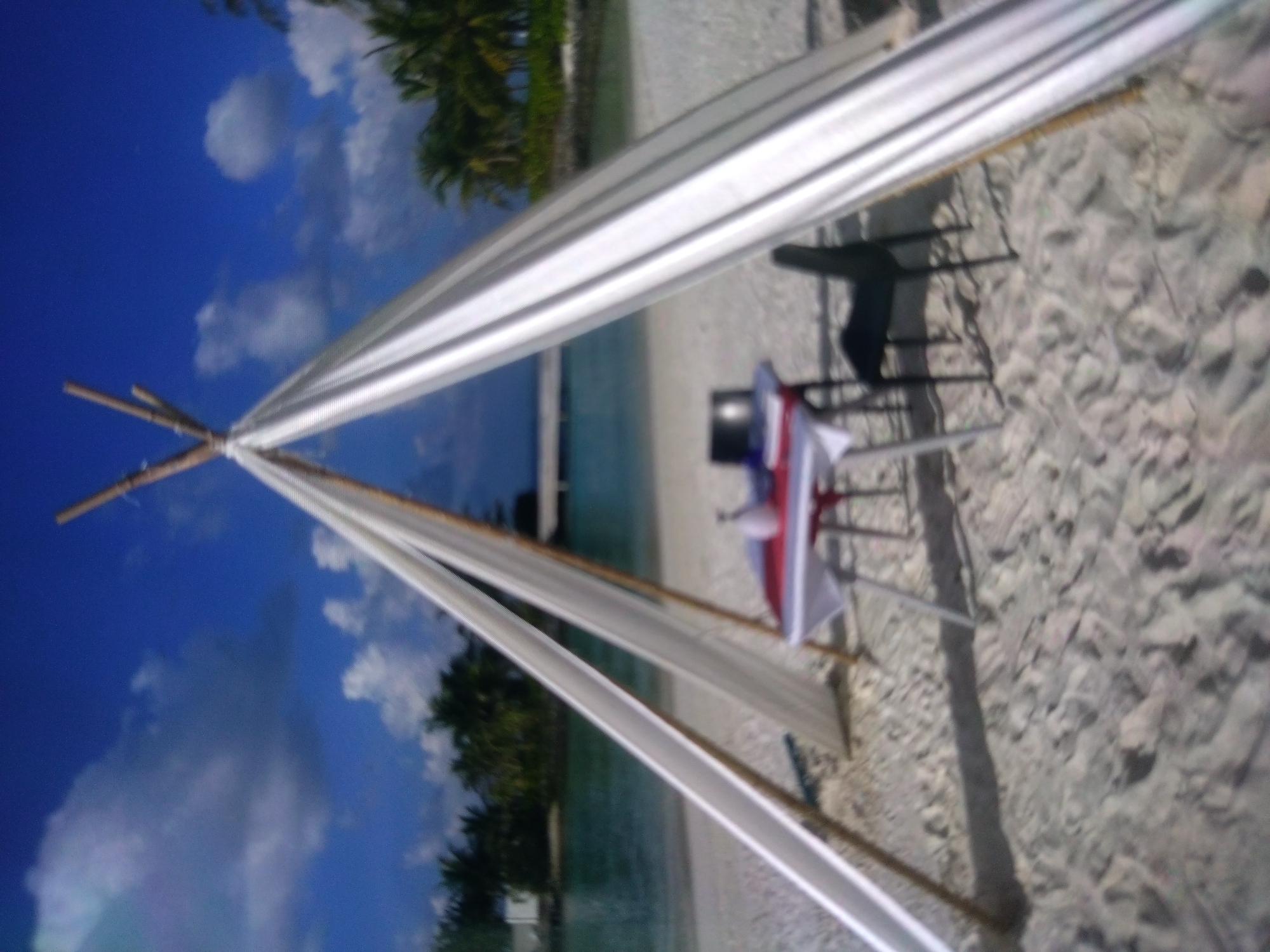 The image depicts a beach wedding setup under a blue sky with scattered clouds, possibly in a tropical or coastal region, not the city of Stara Zagora, Bulgaria. The structure is a simple bamboo frame draped with white fabric, forming a canopy. Under the canopy, there are chairs. The beach is sandy. The ocean is visible in the background.