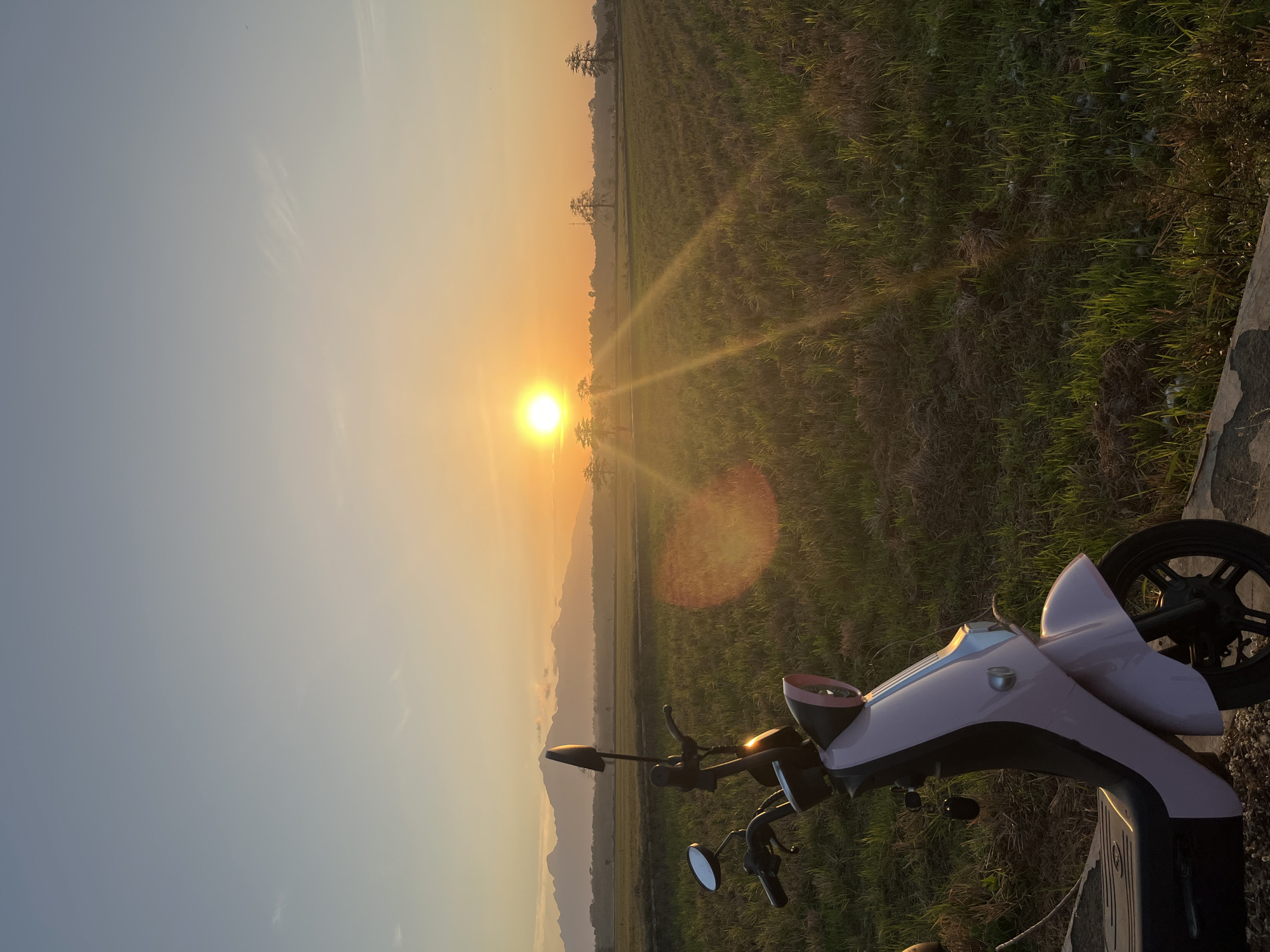 A pink electric scooter is parked in the foreground, facing the right side of the image. The scooter is positioned on a dirt path next to a field of green grass. In the background, a vibrant orange sun is setting over a vast field, casting long shadows and creating a lens flare effect. Distant mountains are visible on the horizon, silhouetted against the colorful sky. The scene is set in the village of Karangrandu, Indonesia. The overall atmosphere is peaceful and serene, with the warm glow of the sunset illuminating the natural landscape.
