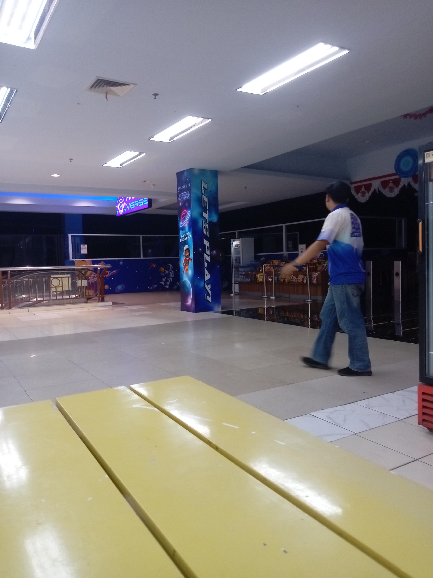 A young man in a white and blue shirt and blue jeans walks across a tiled floor in what appears to be an indoor public space, possibly a mall or entertainment center, in Batam, Indonesia. The man is captured mid-stride, his right arm extended slightly. The environment features fluorescent lighting, ventilation systems on the ceiling, and a decorative pillar with "LET'S PLAY!" graphics and space-themed imagery. In the foreground, a bright yellow table is partially visible. In the background, a railing and a wall mural depicting a starry sky and astronauts can be seen. The overall lighting suggests it is daytime or that the artificial lights are the primary source of illumination.