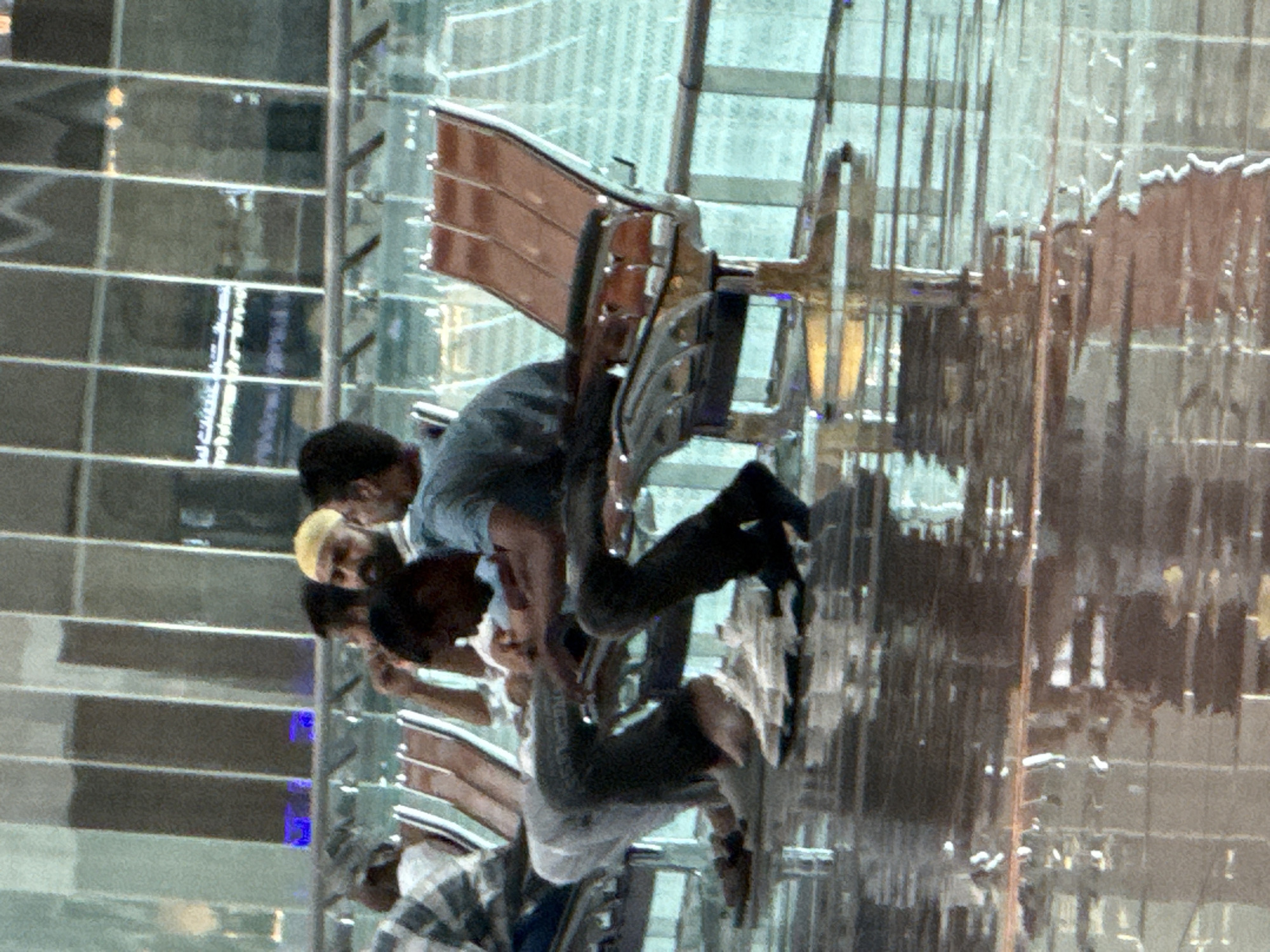 The image shows a group of people seated on brown chairs with metallic bases, likely in an airport lounge or a similar public waiting area. The chairs are arranged in rows. The floor appears to be wet and reflective, suggesting a recent cleaning or spillage. In the background, there is a large glass facade with vertical panels, reflecting lights and possibly the interior of the building. Some of the individuals are wearing casual clothing, and one person is wearing a yellow cap. The overall atmosphere is that of a transit or waiting area. The location is Muscat International Airport in Seeb Province, Oman.