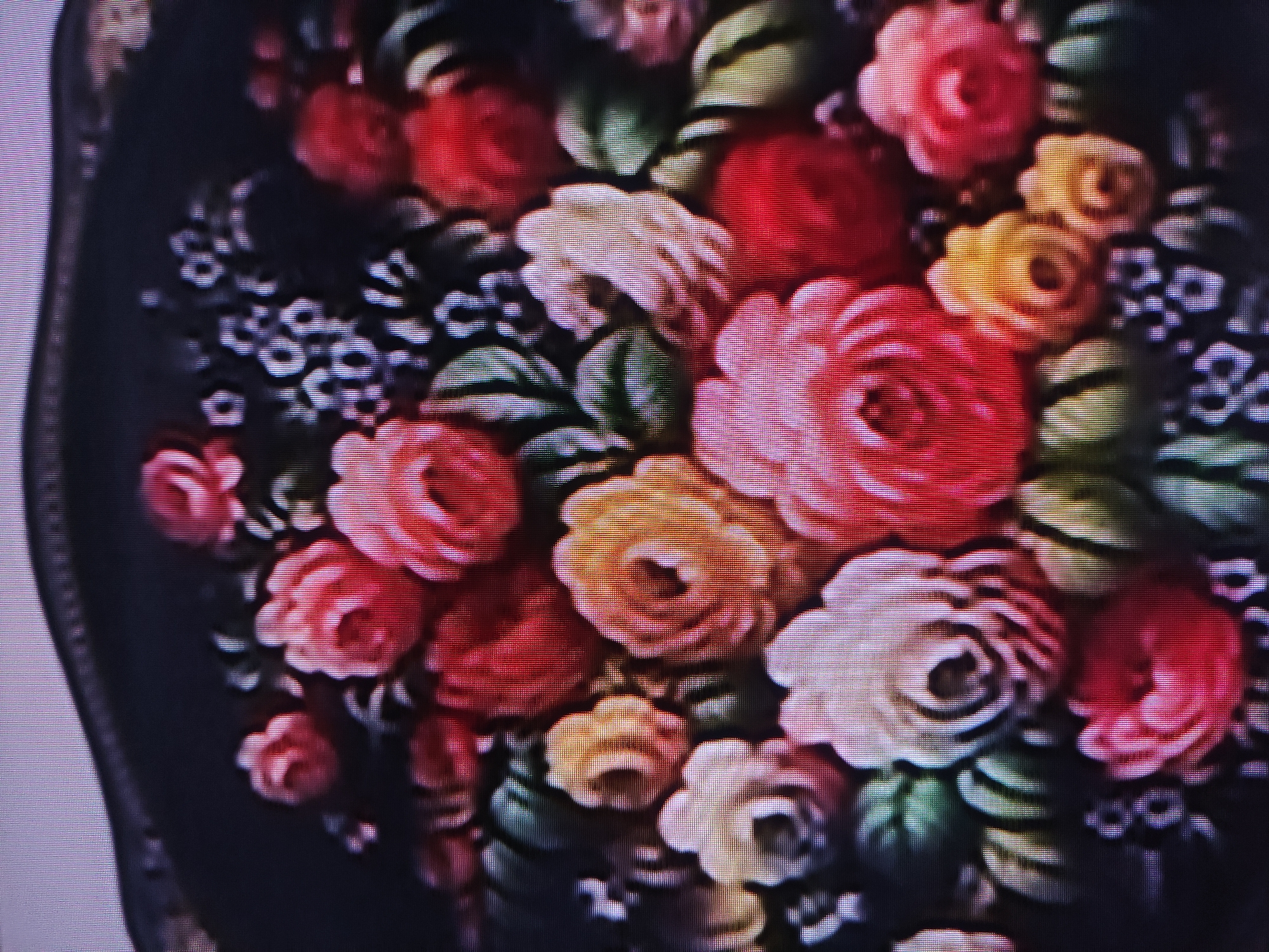 The image shows a close-up of a floral painting, likely on a dark surface, possibly a tray or piece of furniture. The painting consists of various flowers, predominantly roses, in shades of red, pink, yellow, and white. Green leaves are interspersed among the flowers. There are also small blue or purple flowers scattered throughout the bouquet. The overall style is reminiscent of traditional folk art or floral decorative painting. There is no information or evidence that places the location of this image in Pavlodar, Kazakhstan. The image contains no people, subjects, objects, actions, interactions, or descriptions of an environment that might lend itself to an accurate forensic interpretation.