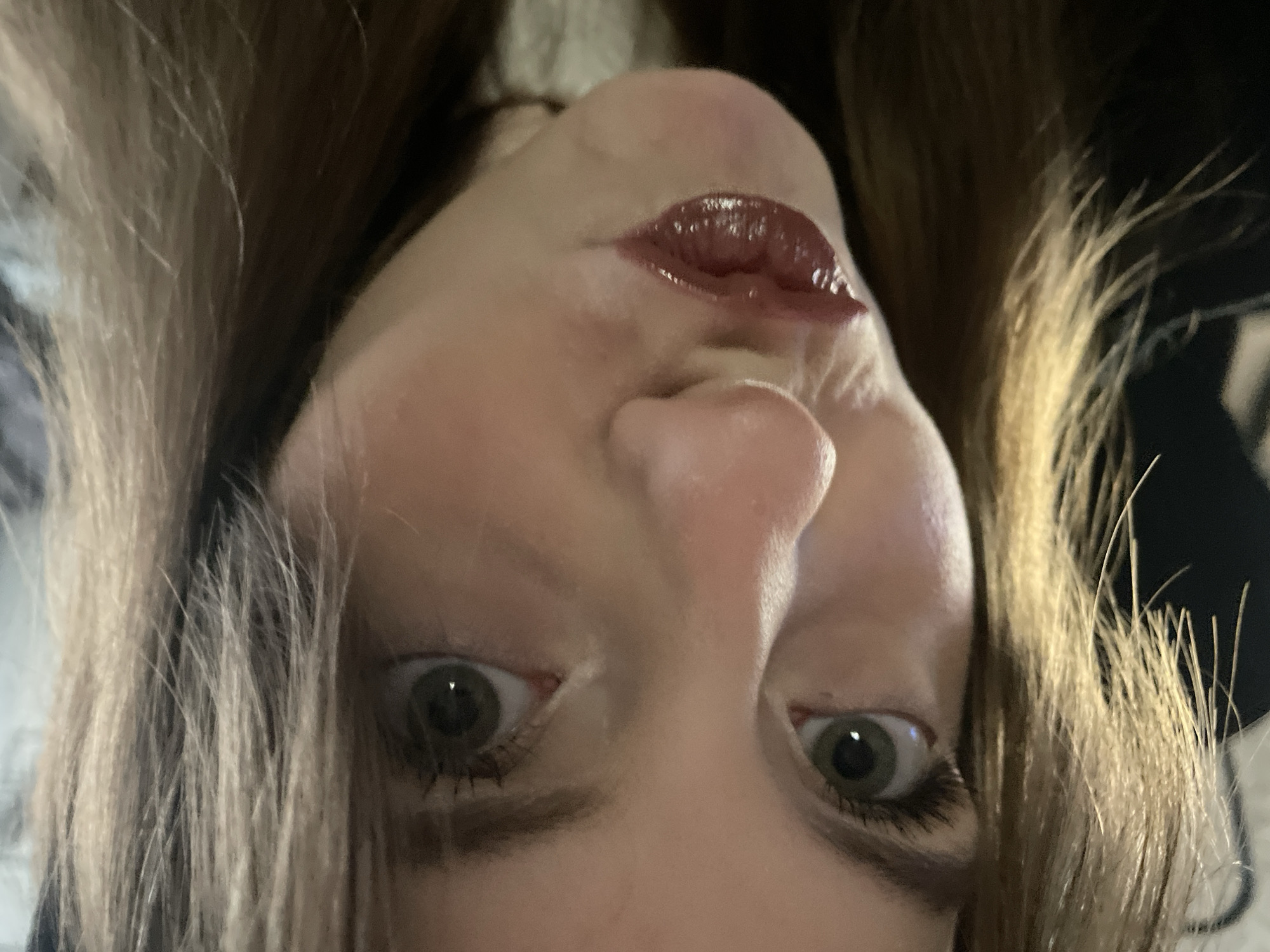 The image is a close-up, inverted selfie of a woman. Her face is centered, with her eyes looking directly at the camera. She has fair skin, light brown hair, and dark-colored eyes. Her lips are coated with a shiny, dark red lip gloss. The background is out of focus and appears to be dark. There is no information to suggest this image was taken in Фоки, Russia.