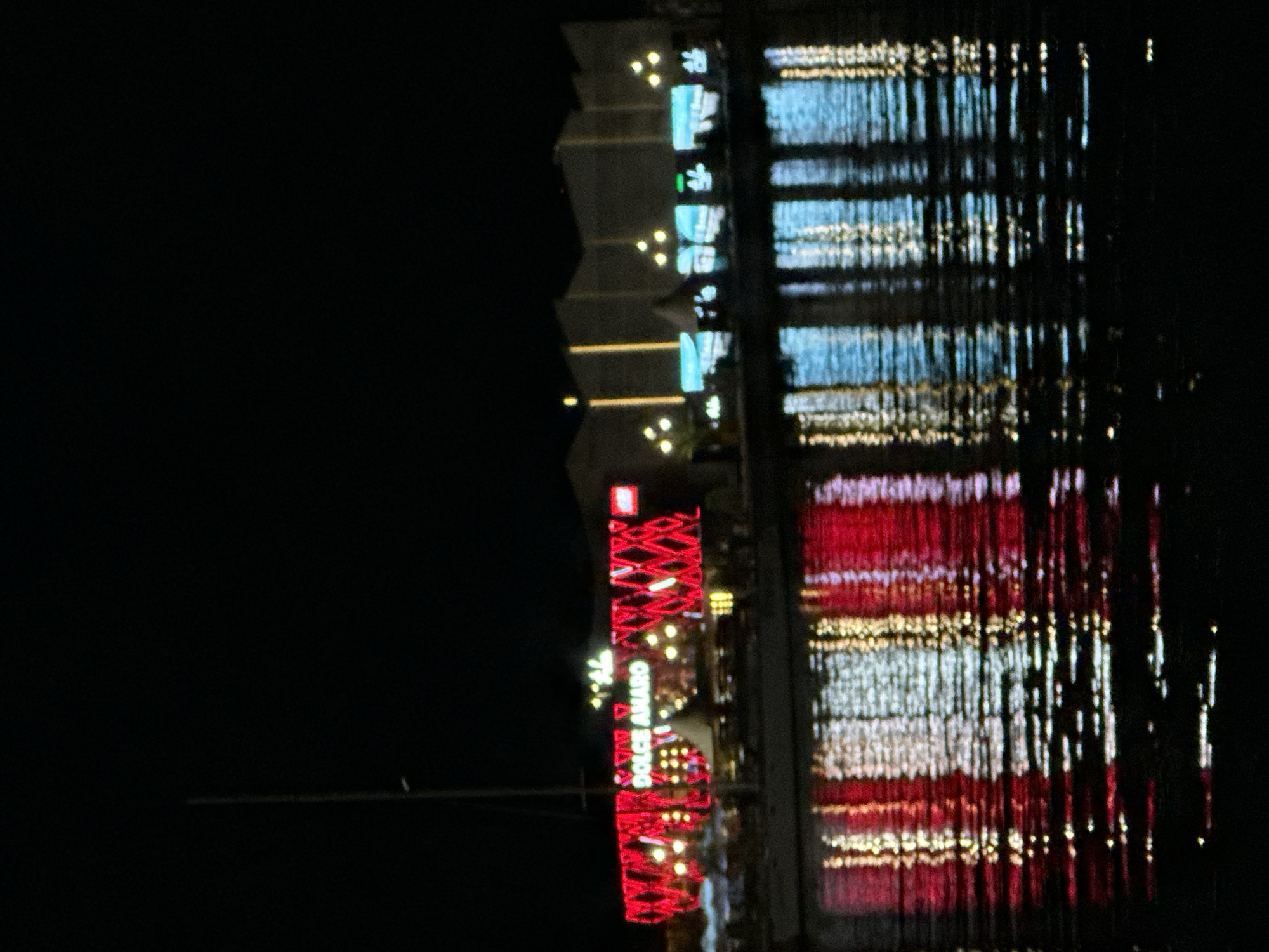 The image shows a nighttime scene in Varna, Bulgaria, reflected in water. A building is visible, illuminated with lights. A red column-like structure with a criss-cross pattern of lights is labeled with "DOLCE AMARO". The rest of the building has different levels of illuminated text, as well as string lights. The reflection in the water creates distorted, elongated streaks of light. The scene appears to be photographed at night, as the background is dark.