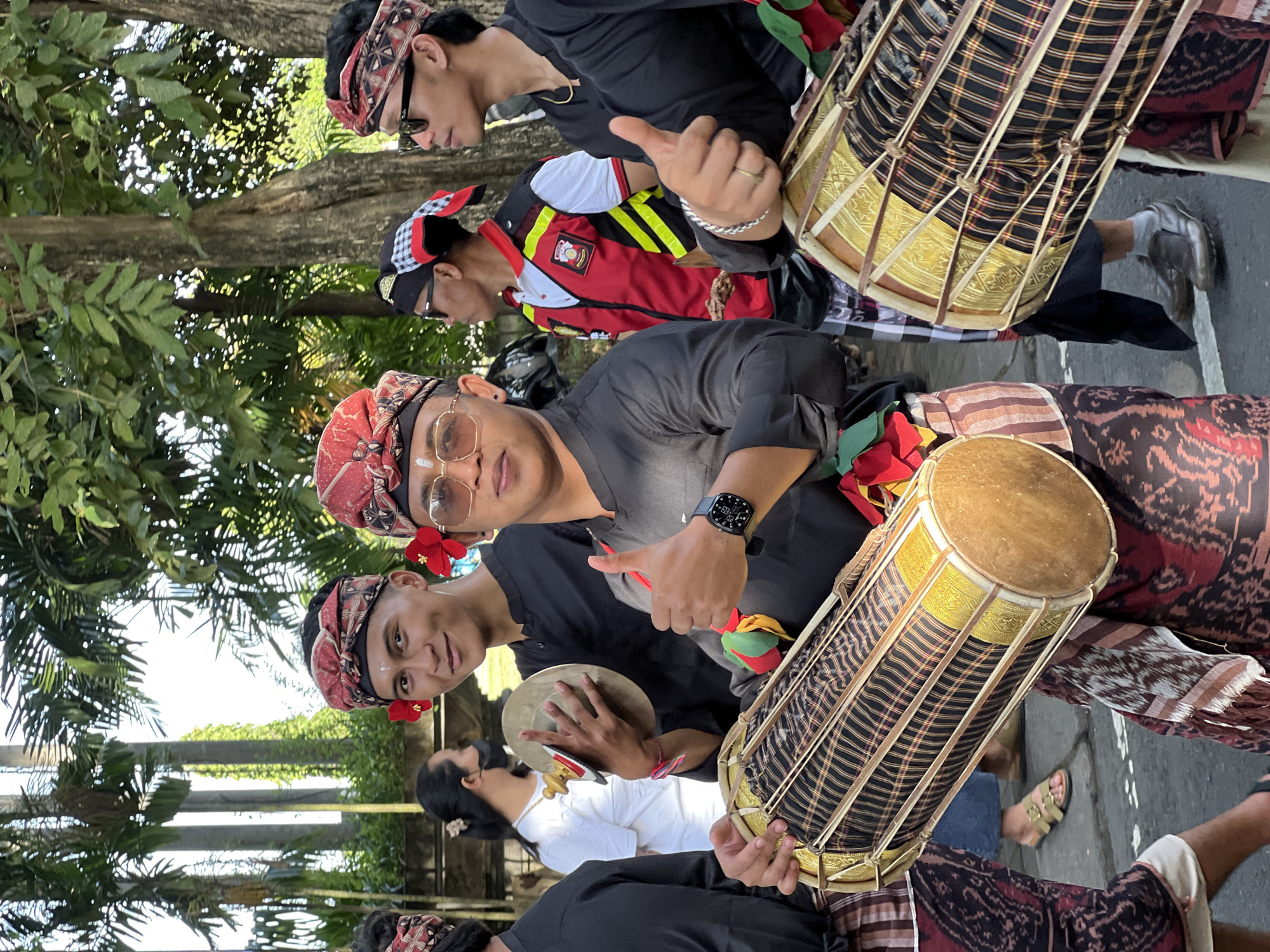 In Denpasar, Indonesia, a group of young men in traditional Balinese attire participate in a street procession. The men are wearing black long-sleeved shirts, patterned sarongs, and red and black headbands adorned with red flowers. Two of the men are holding cylindrical drums, which are decorated with intricate patterns. One of the men is playing a cymbal, and another is giving a thumbs-up gesture. In the background, palm trees and lush foliage create a tropical atmosphere. A woman in a white shirt is visible in the mid-ground, while other individuals in various attire are also present, suggesting a festive or ceremonial event. The scene appears to be taking place outdoors during daylight.