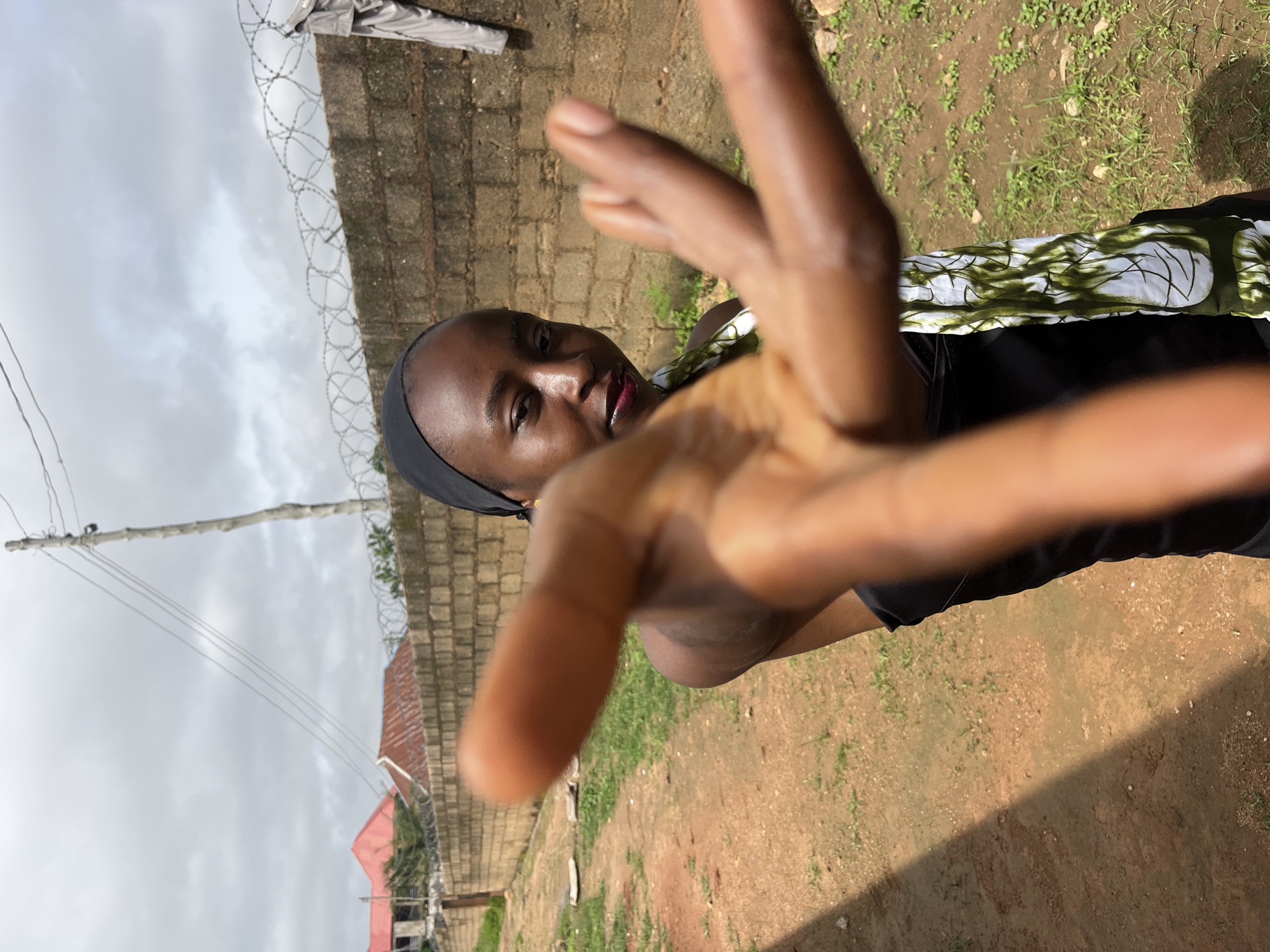 A person, likely a young woman, is captured in a close-up shot with her hand reaching towards the camera, obscuring a significant portion of the frame. She is wearing a black head covering and a black garment with a patterned white and green fabric across her chest. Her gaze is directed at the camera, and she appears to be smirking or smiling slightly, with visible lipstick.
The background suggests an outdoor setting in Bukuru, Nigeria. A brick wall, topped with barbed wire, forms a boundary behind the subject. Beyond the wall, a red-roofed building and some trees are visible, indicating a residential or semi-urban area. Power lines stretch across the sky, and the ground appears to be dirt with scattered patches of grass. The sky is overcast, suggesting it might be a cloudy day, possibly late afternoon due to the angle of the shadows. The overall mood seems casual and personal, as if the person is posing for a selfie or interacting directly with the photographer. No visible text is present in the image.