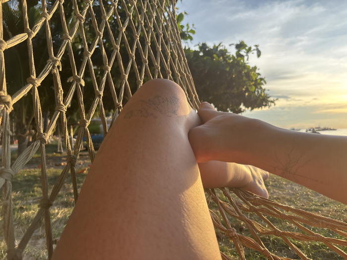 The image depicts a first-person perspective from an individual resting in a light brown woven net hammock in Koh Tao, Thailand, facing towards the sea during either sunrise or sunset. The foreground features the individual's bare legs and right arm. The left leg is bent, displaying a faint, faded linear tattoo on the upper thigh. The right forearm, resting across the left leg, exhibits a delicate, branching tattoo resembling a tree or nerve structure. The hammock's knotted cord netting, forming diamond patterns, is prominent on the left and bottom. Beyond the hammock, a grassy area with palm trees and tropical foliage is visible. The background shows the sea with multiple vessels silhouetted against a brilliant golden sky, characteristic of low sun angle. No other individuals are present.