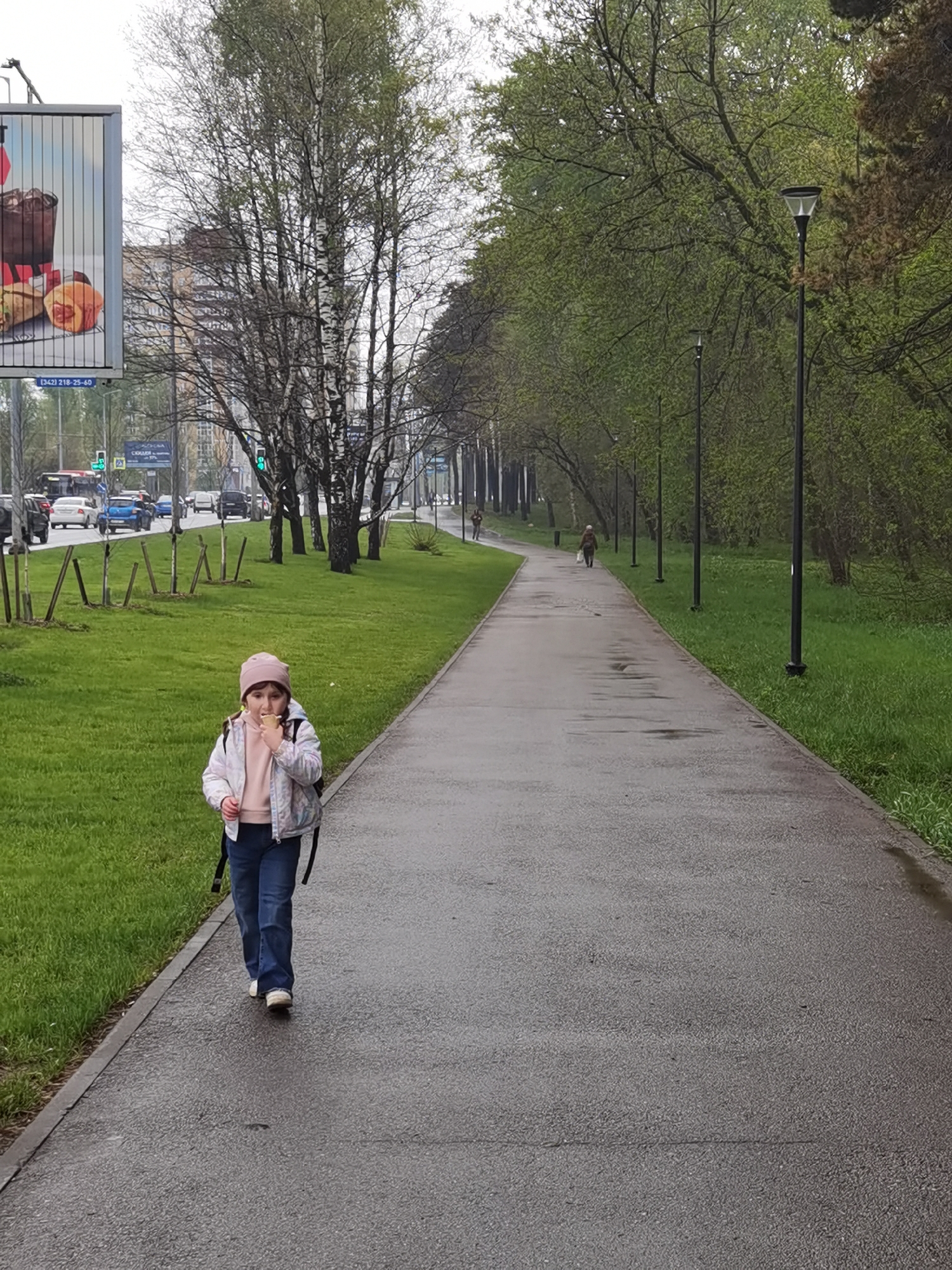 A young girl with a pink hat, jacket, and jeans is walking along a wet sidewalk, eating a snack. A backpack hangs on her back. The sidewalk stretches into the distance, flanked by green grass on both sides and trees lining the path. In the background, other pedestrians are visible, and lampposts stand along the sidewalk. Further in the background, buildings and cars are visible, as well as a billboard. The location is in the city of Perm, Russia.