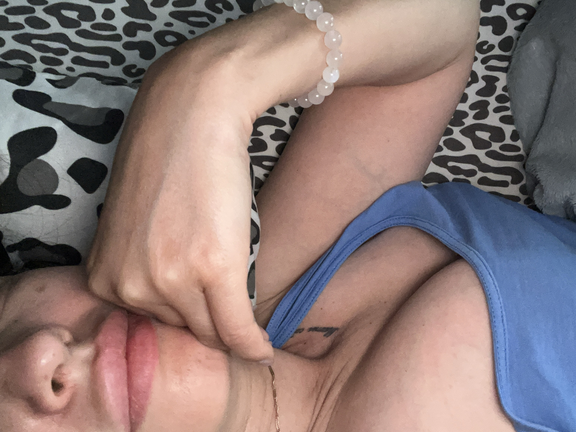 The image is a close-up, overhead shot of a person lying down. The person's face is partially visible, with their lips and nose in focus. Their eyes are closed and their hand is gently touching their face. They are wearing a blue tank top and a beaded bracelet on their wrist. The background is a leopard-print fabric and a gray blanket. The lighting is soft and diffused, suggesting an indoor setting during the daytime. The overall mood appears relaxed and serene. There is a small tattoo visible on the person's chest, but no text is discernible.