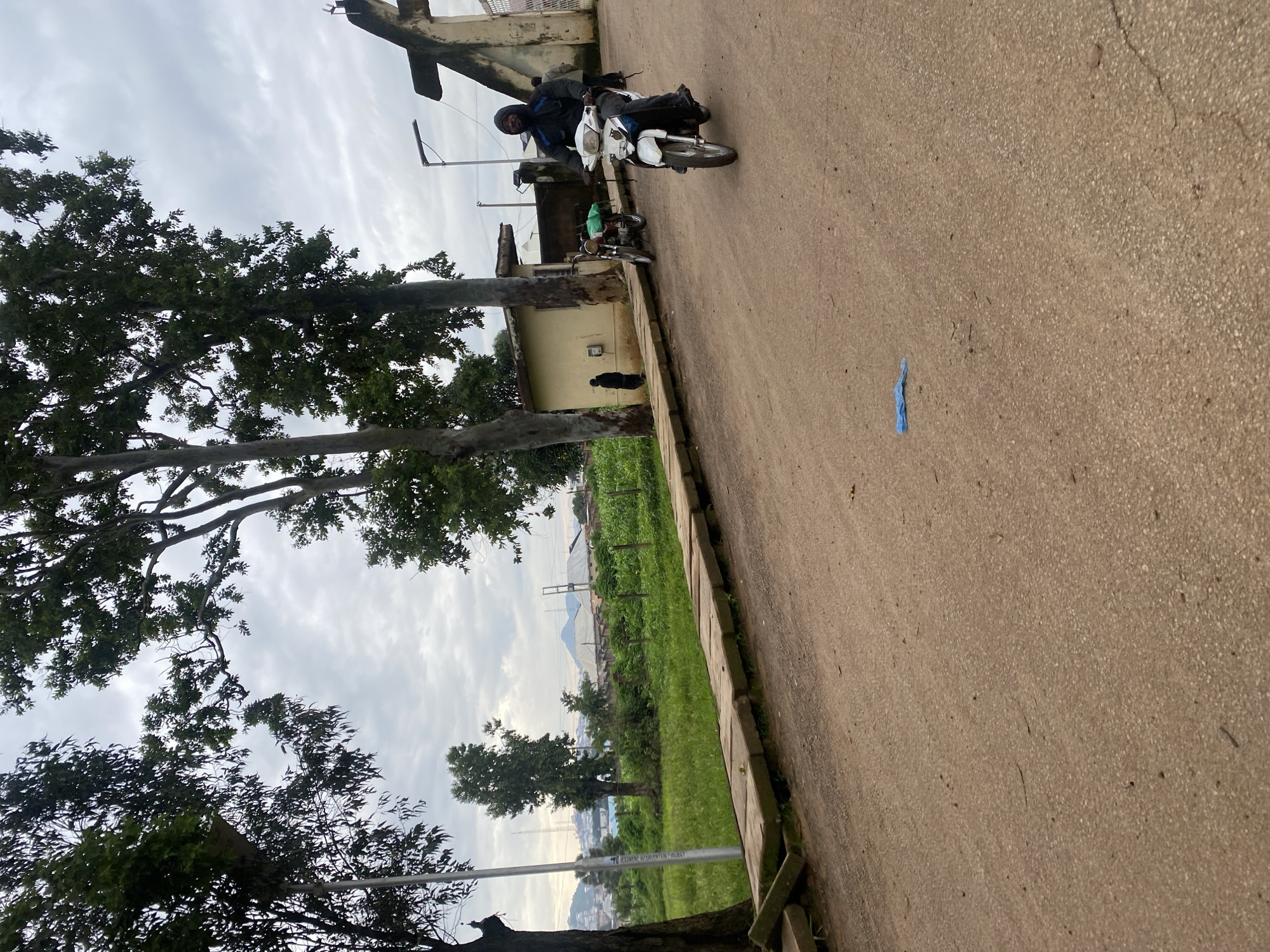 In Rafin Par, Nigeria, a person wearing a dark hooded jacket rides a white motorcycle on a dirt road. The motorcycle has a green bag strapped to its rear. Two other motorcycles are parked nearby, one red and one red and black. Behind them is a yellow building and a concrete archway. A person in black stands near the yellow building, and two people are visible near the archway. In the background, there is lush green vegetation, including trees and a field, and a cityscape with buildings and hills under a cloudy sky. A light pole is visible on the right side of the image. A blue plastic bag is on the dirt road in the foreground.