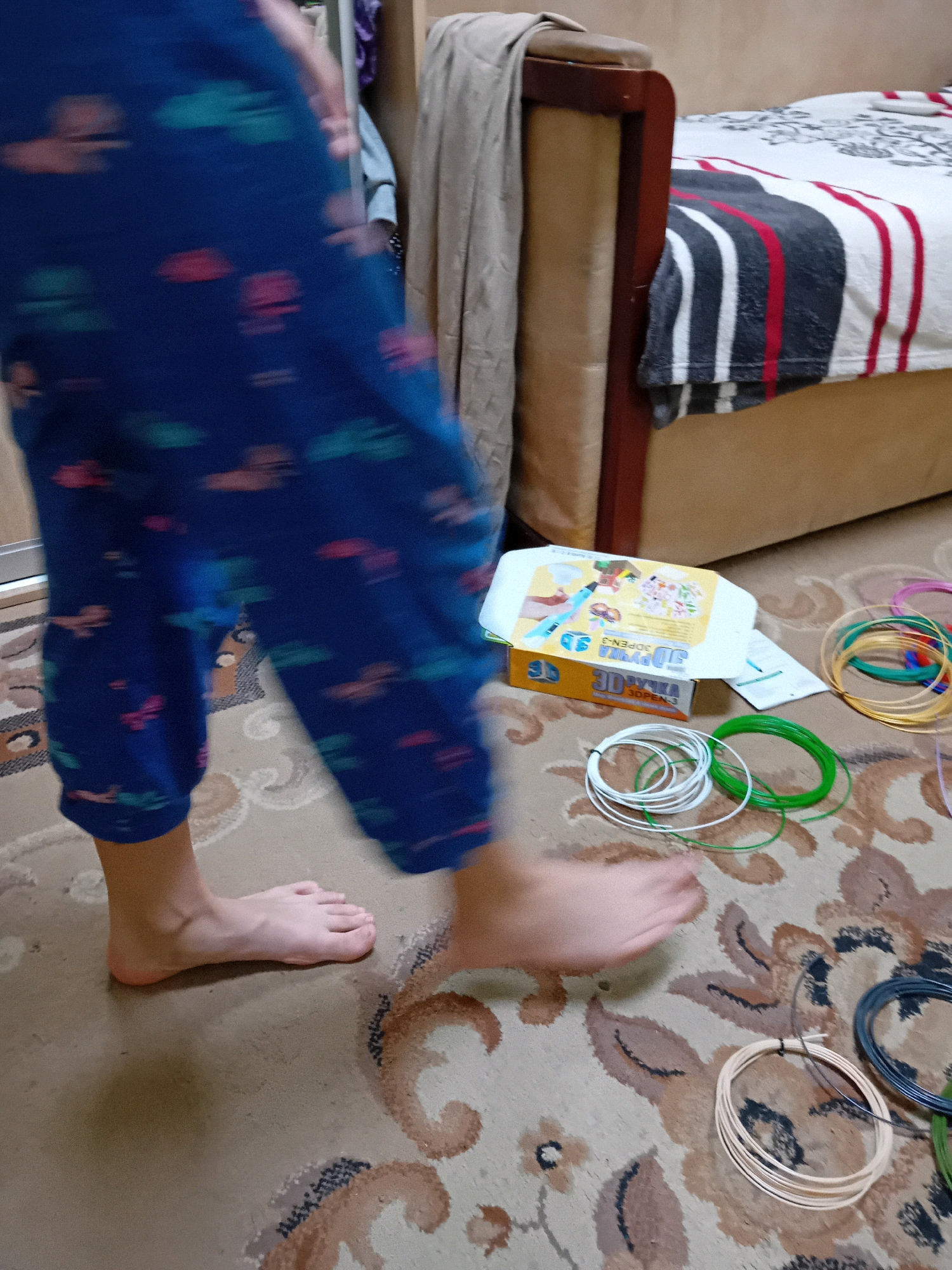 The image depicts an indoor scene, likely a residential room, possibly in Chuhuiv, Ukraine, given the context. In the foreground, a person's lower legs and feet are visible, wearing blue pajamas with a pattern of colorful shapes. The person's feet are bare and appear to be in motion, suggesting they are walking. The floor is covered with a patterned rug. Scattered on the rug are several spools of filament, predominantly in white, green, gold, and black, indicative of supplies for a 3D pen. To the right, a box for a 3D pen is partially visible, along with other related items. In the background, a bed with striped bedding and a headboard is present, along with a beige upholstered piece of furniture and a draped cloth. The lighting suggests an indoor setting, possibly daytime.