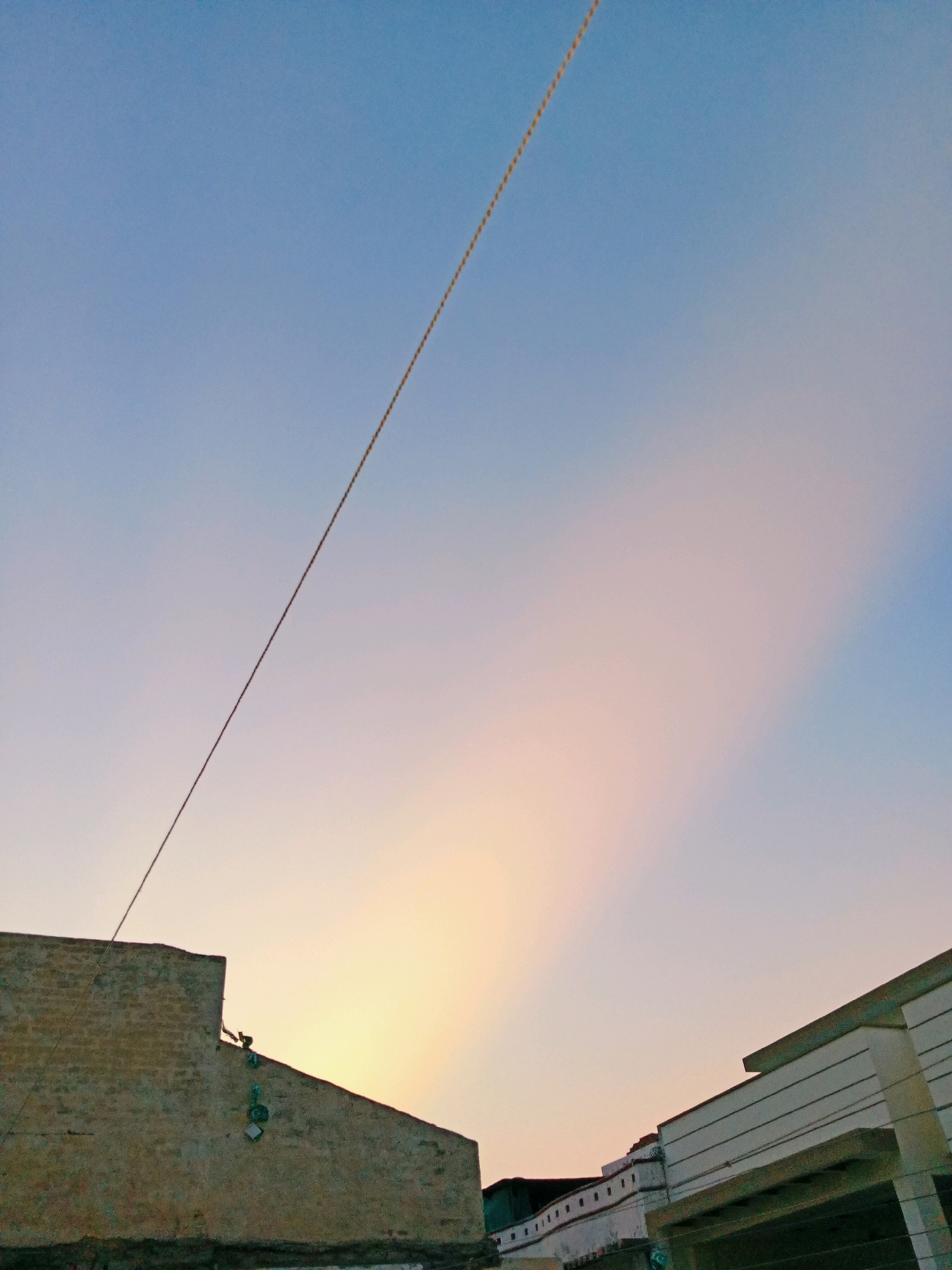A daytime image captures a sky with subtle pastel hues transitioning from a pale blue at the top to warmer yellow and pink tones near the horizon. A thin, dark utility line diagonally bisects the frame from the upper left to the lower right. In the lower portion of the image, the rooftops of two distinct buildings are visible. The building on the left is made of rough, weathered concrete or brick and features a sharp, angled roofline. Attached to its slanted surface are several green and white square-shaped objects, possibly decorative elements or attached lights. The building on the right has a more modern facade, with clean lines and horizontal white siding. The overall scene suggests an urban or semi-urban environment, likely within Bahawalpur City Tehsil, Pakistan, during a time of day when the sun is low, creating the soft sky colors. There are no people or animals visibly present in the frame.
