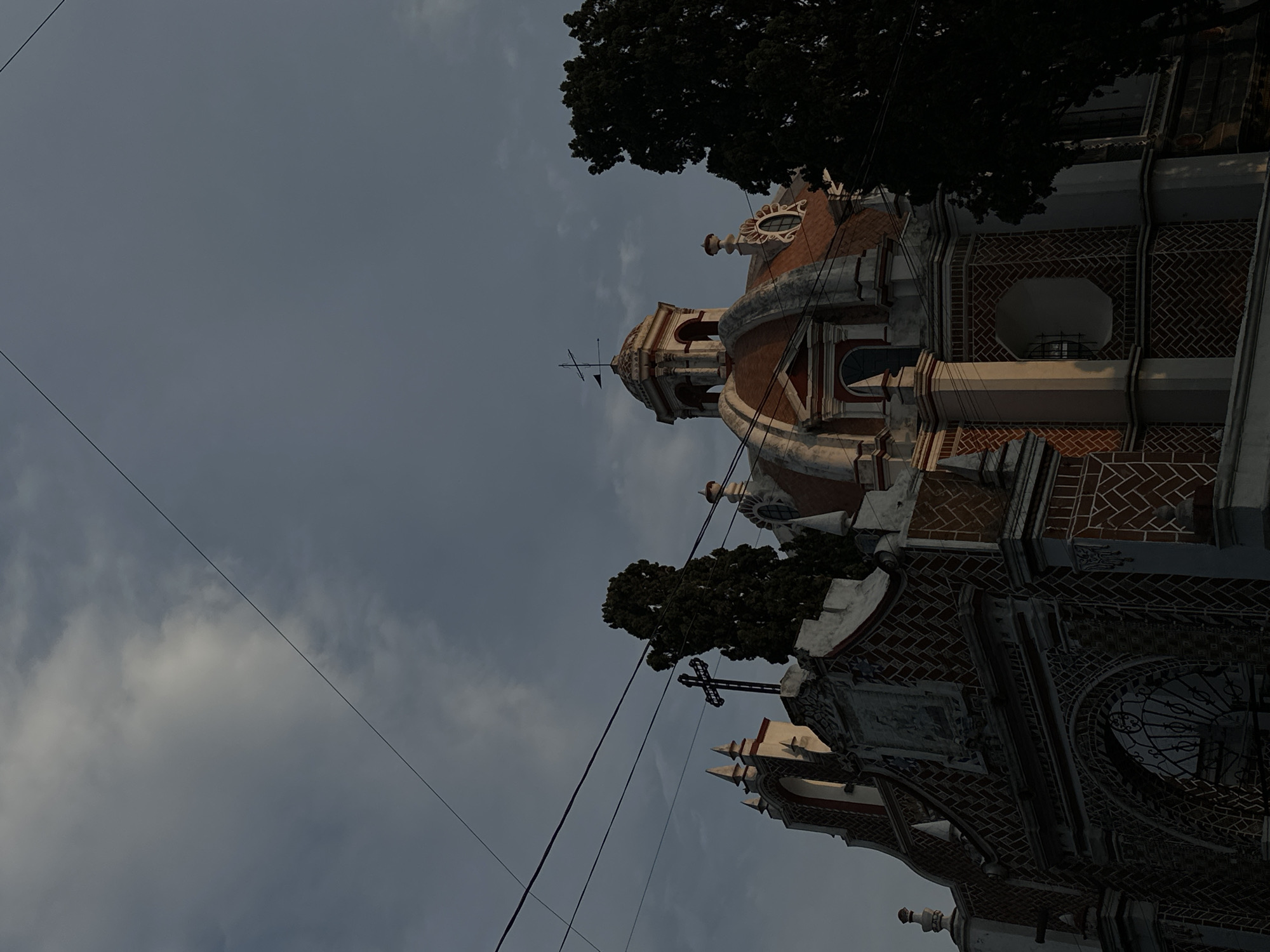 The image shows a low-angle view of a church in Unknown, Unknown. The church features a dome with a cross on top, and brickwork patterns on the exterior walls. Wires are visible against the gray sky. Trees are positioned on either side of the church.