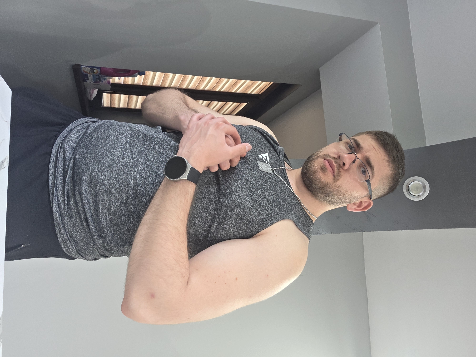 A man with a beard and glasses is photographed indoors from a low angle, appearing to stand. He is wearing a gray tank top, black shorts, and a smartwatch. He has a silver necklace. The background includes gray walls and ceiling, a light fixture, and a small recessed window with blinds. The location is Yagodovo, Bulgaria cannot be determined from the image.