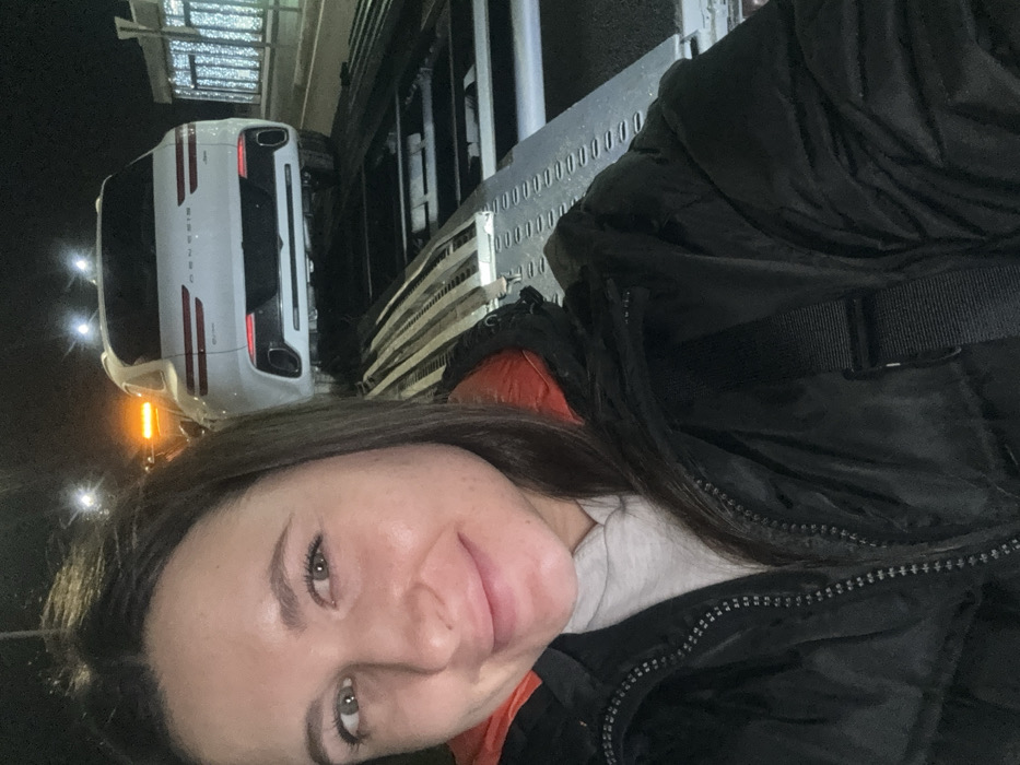 A woman with brown hair and fair skin smiles at the camera. She is wearing a dark puffer jacket. Behind her is a car carrier trailer with a white SUV parked on it. The lighting suggests it is nighttime or a dimly lit area.