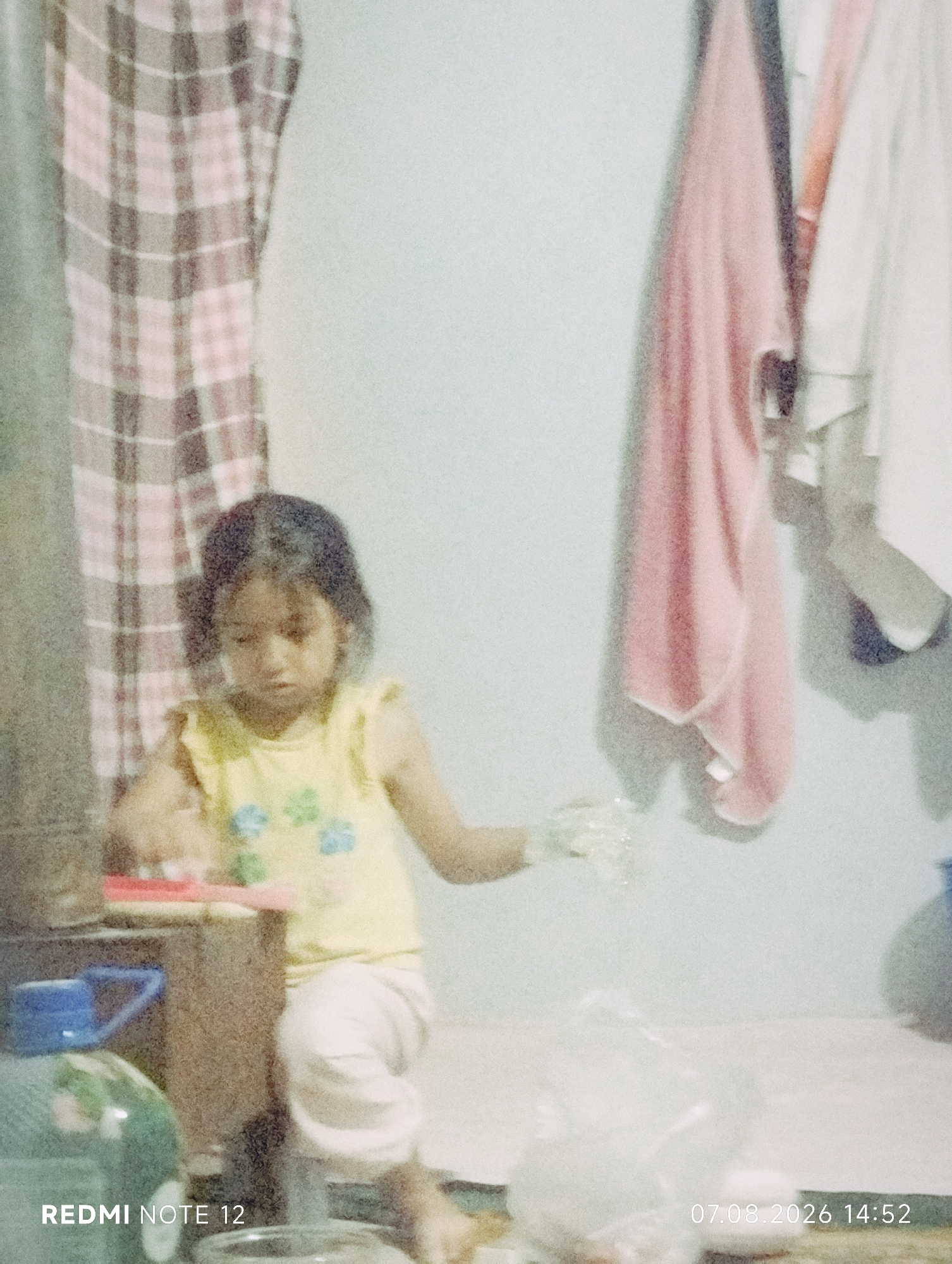 A young girl with dark hair and a yellow top with floral patterns is standing indoors. She is wearing white pants. She is reaching out with her right hand, which appears to be holding something clear and bubbly, possibly soap or water. Her left hand is on a wooden surface. Behind her, to the left, is a checkered curtain or fabric in shades of pink and brown. To the right of the girl, several items of clothing are hanging on a wall, including a pink towel and a white garment. A large green plastic bottle with a blue cap is visible in the foreground on the left. A plastic bag is on the floor to the right of the girl. The lighting suggests it is daytime, and the scene appears to be in a domestic setting. The bottom of the image shows the text "REDMI NOTE 12" and a date and time stamp, "07.08.2026 14:52".