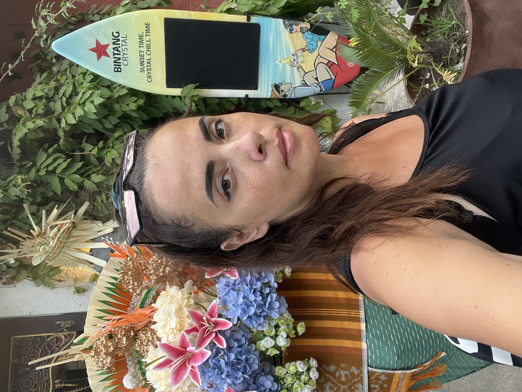 In Kuta, Indonesia, a woman stands in front of a decorative display and a Bintang beer surfboard. The woman, with dark brown hair pulled back and sunglasses perched on her head, is wearing a black tank top. To her left is a colorful arrangement of artificial flowers, featuring pink lilies, blue hydrangeas, and cream-colored blossoms, accented with orange and green decorative elements. The background is a wall covered in green foliage. To her right is a surfboard-shaped sign for Bintang Crystal, with the text "SUNSET TIME, CRYSTAL CHILL TIME" and a depiction of a couple on a beach. A potted palm is visible at the base of the surfboard.