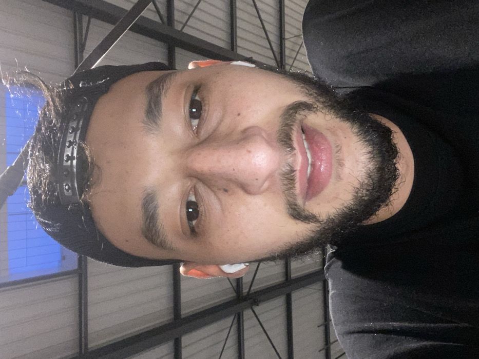 A man with a beard, dark hair, and dark eyes faces the camera. He wears a black beanie with a silver-studded band and has a white wireless earbud in his right ear. He wears a black shirt, and the background features an industrial ceiling with exposed metal beams and a window with blue and white light. The image is vertically oriented.
