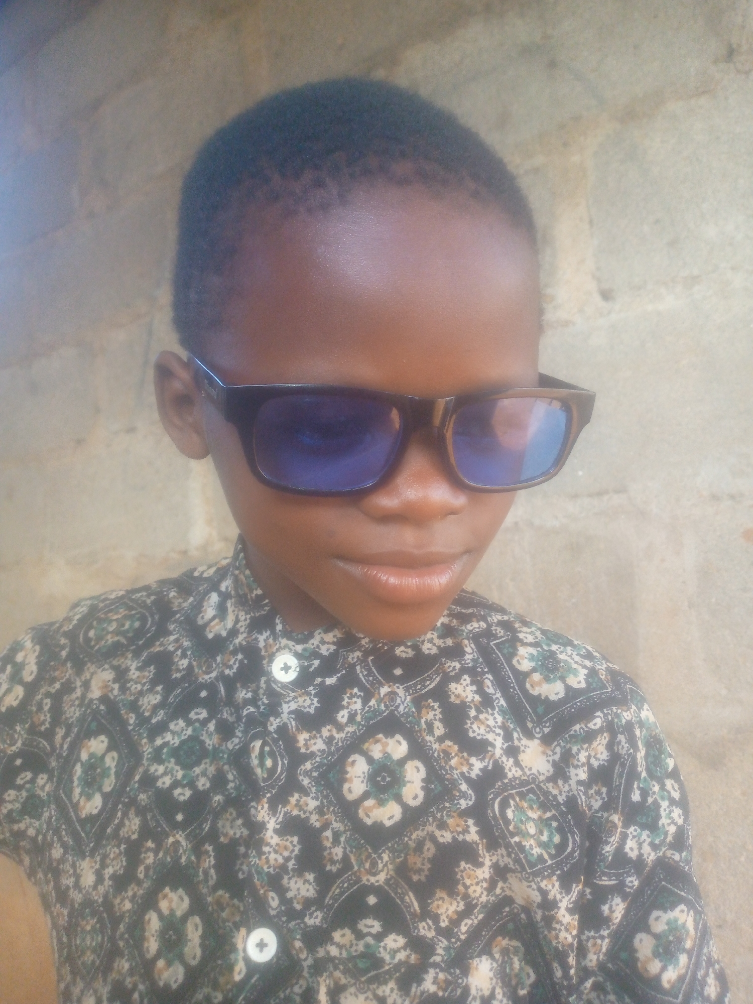 A close-up portrait features a young person with dark skin and short, dark hair, wearing sunglasses. The individual faces forward, with their head slightly tilted downward, and their eyes are obscured by large, dark-framed sunglasses with blue-tinted, reflective lenses. A slight smile is visible on their lips.
The subject is wearing a collared shirt with an intricate, repeating pattern. The shirt's base color appears dark, possibly black or dark blue, adorned with lighter designs in shades of white, cream, and green, featuring floral or geometric motifs. Two white, four-holed buttons are visible on the front of the shirt.
The background consists of a light-colored, textured wall, possibly concrete blocks or plaster, suggesting an outdoor or semi-outdoor setting. The lighting is bright and appears even, indicating a daytime photograph. The person is posing for the camera. No discernible text is visible in the image.