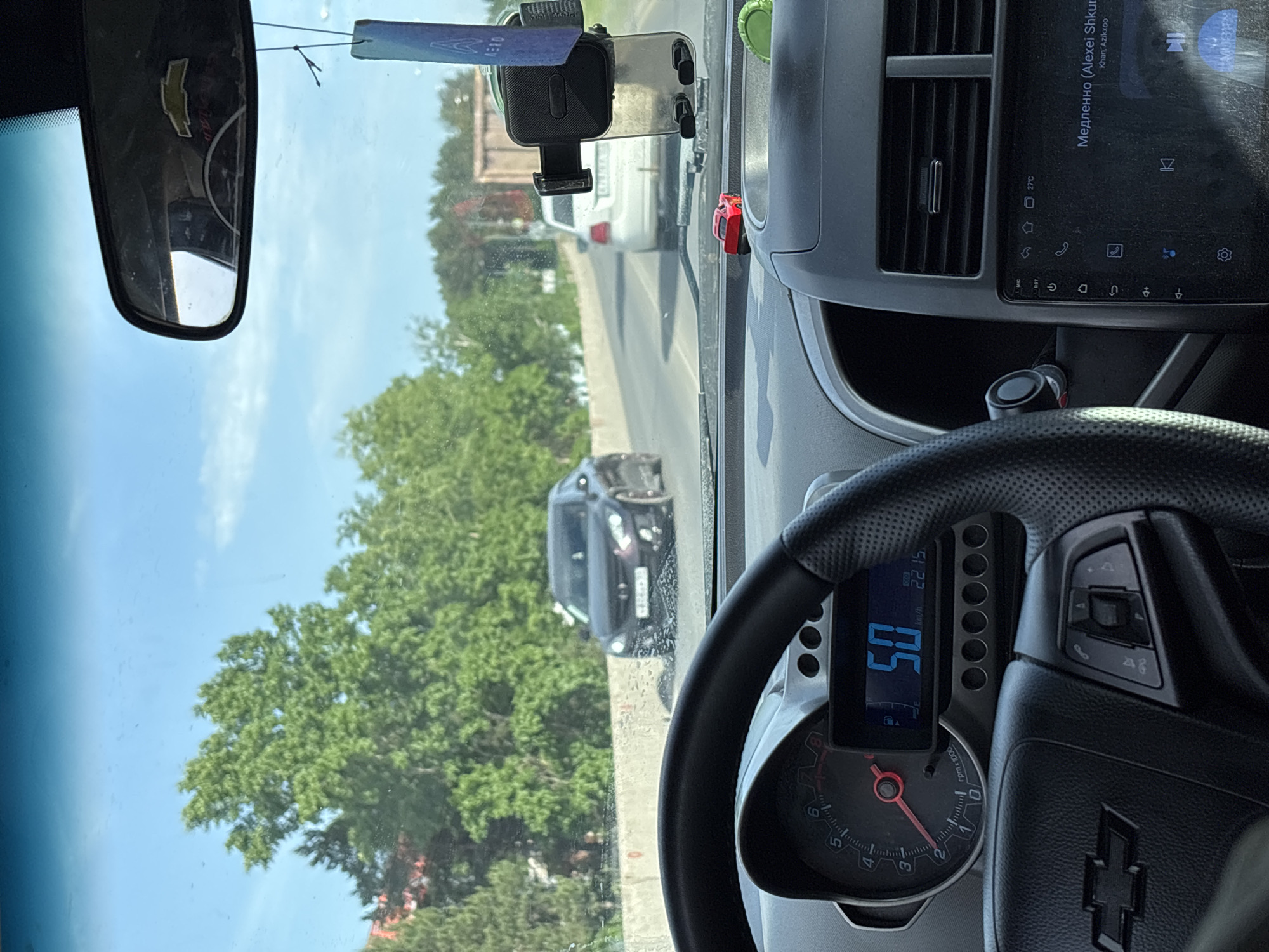 The image is an interior view from the driver's seat of a car, likely a Chevrolet, as indicated by the steering wheel emblem and the rear-view mirror. The speedometer reads 50 km/h, and the tachometer needle is just below 3. A digital display shows additional car information. The car's infotainment system displays a music track by Alexei Shkurko.
Outside, the car is on a road with other vehicles present. The environment includes trees and greenery. Given the OCR text identifying the Cyrillic alphabet, it can be safely assumed that this location is Chaikovsky, Russia. The camera is positioned inside of the car and captures a forward-looking view, including surrounding vehicles and foliage.