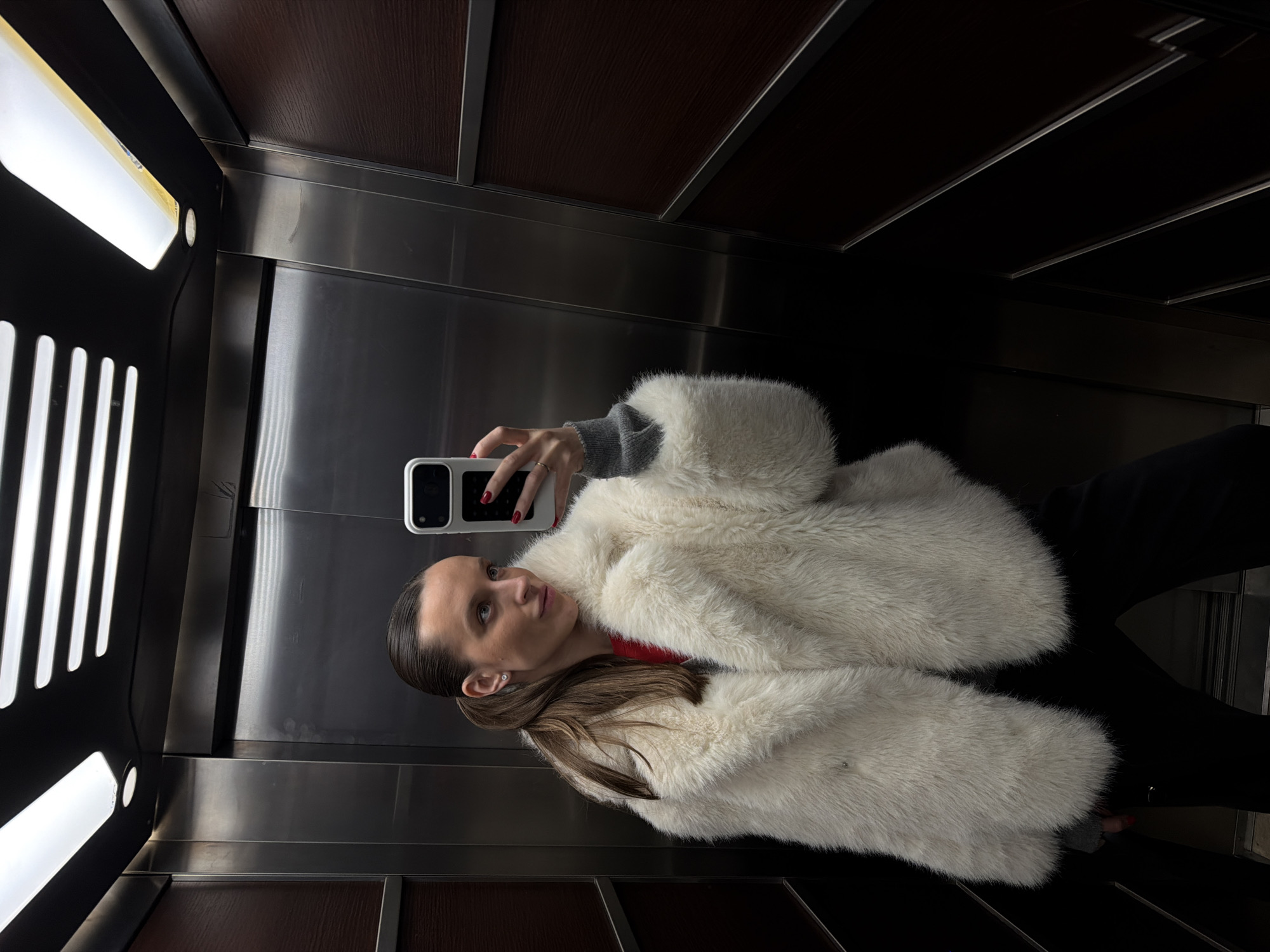 A woman is taking a selfie in an elevator. The elevator's interior features stainless steel walls and a dark brown ceiling and base trim. A white, fur-like coat is worn over a gray long-sleeved garment, visible at the cuffs. The woman's hair is pulled back. The camera is pointed towards her. The photo does not provide location information for Budva, Montenegro.