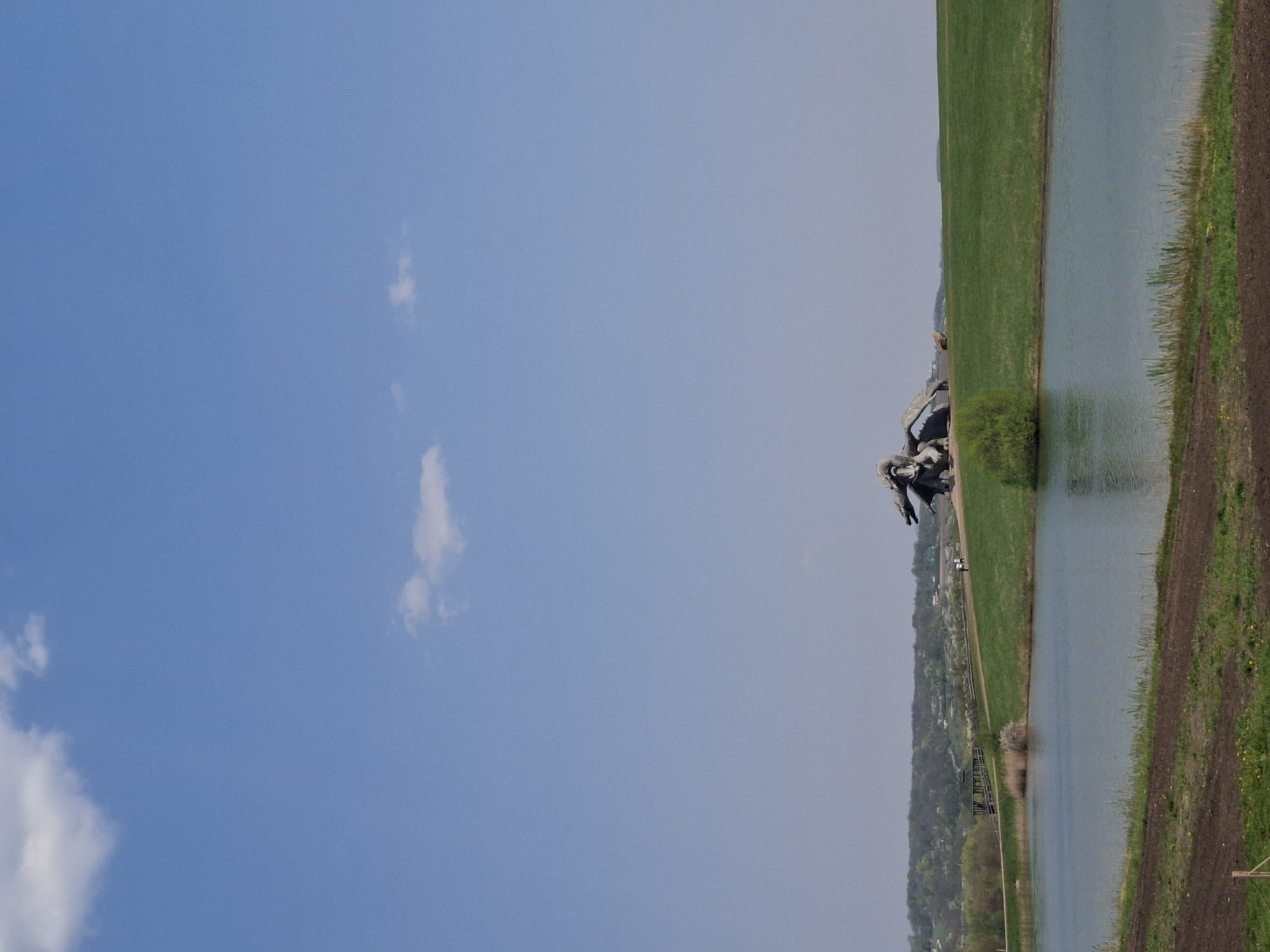 The image depicts a landscape adjacent to a body of water, likely a river or lake. The sky is blue with scattered clouds. Alongside the water, there's a green grassy area and tilled earth. In the background, there's a tree line. A sculpture or monument is visible in the foreground. This image seems to be in Алисово, Russia.