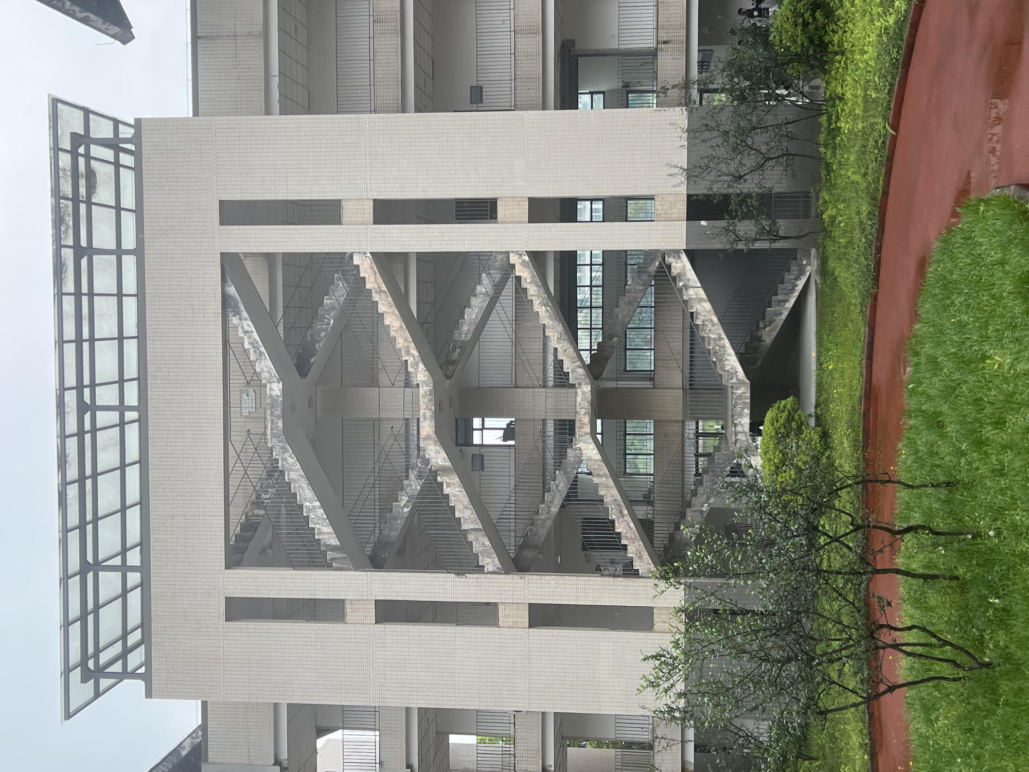 The image shows the exterior of a building, likely a school, with an exposed staircase. The building's facade is light-colored tile. The staircase is made of concrete and metal railings. There is greenery, including trees and grass, surrounding the building. A red running track is visible to the right. It is raining. The picture was taken at Jiangjin, China. There are people visible at the upper side of the building.