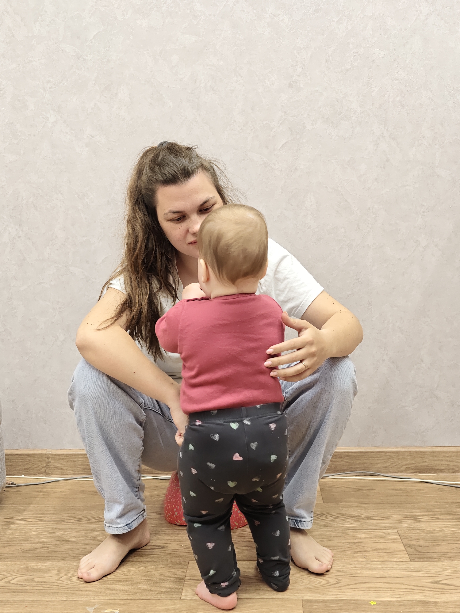 The image depicts an intimate indoor scene featuring a woman and a baby, likely a mother and child, engaging in a tender moment in a room located in Chaikovsky, Russia.
The woman, with long, dark brown hair pulled back in a ponytail, is seen from the front. She wears a white t-shirt and light blue jeans, and is barefoot. She is crouched down on the floor, directly facing the baby, with her gaze intently fixed on the child. Her left hand is gently placed on the baby's back, suggesting support or affection, while her right hand is near the baby's side. Her expression is one of focused attention and care.
The baby, seen from the back, is wearing a red long-sleeved top and dark gray leggings adorned with small, colorful heart patterns. The baby is also barefoot and stands facing the woman, appearing to be in an upright position, possibly practicing standing or taking first steps with the woman's support. A small red object, possibly a cushion or toy, is partially visible under the baby.
The background consists of a light-colored, textured wall. The floor is made of light brown wooden or laminate material. Some white and gray electrical wires are visible along the base of the wall on the floor. The overall ambiance suggests a quiet, domestic setting during what appears to be daytime due to the clear indoor lighting. There is no visible text in the image.