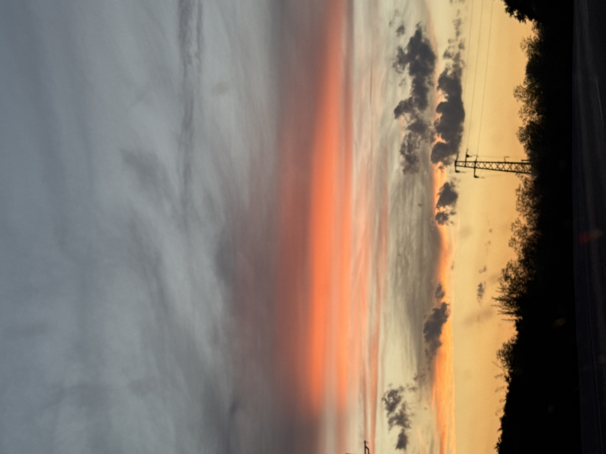 The image captures a sunset over a landscape, likely in or near Veselina, Bulgaria. The sky is a dramatic display of color, with a band of bright orange clouds stretching horizontally across the frame. Above this fiery band, the clouds transition to a gradient of grays, suggesting an approaching storm or the lingering darkness of twilight. Below the orange glow, the sky is a pale yellow, fading into the dark silhouette of trees and vegetation that form the horizon line. A prominent power line tower stands tall against this backdrop, its lattice structure clearly visible. The foreground shows a dark road, hinting that the photograph was taken from a moving vehicle. The overall scene evokes a sense of natural beauty and the transition from day to night.