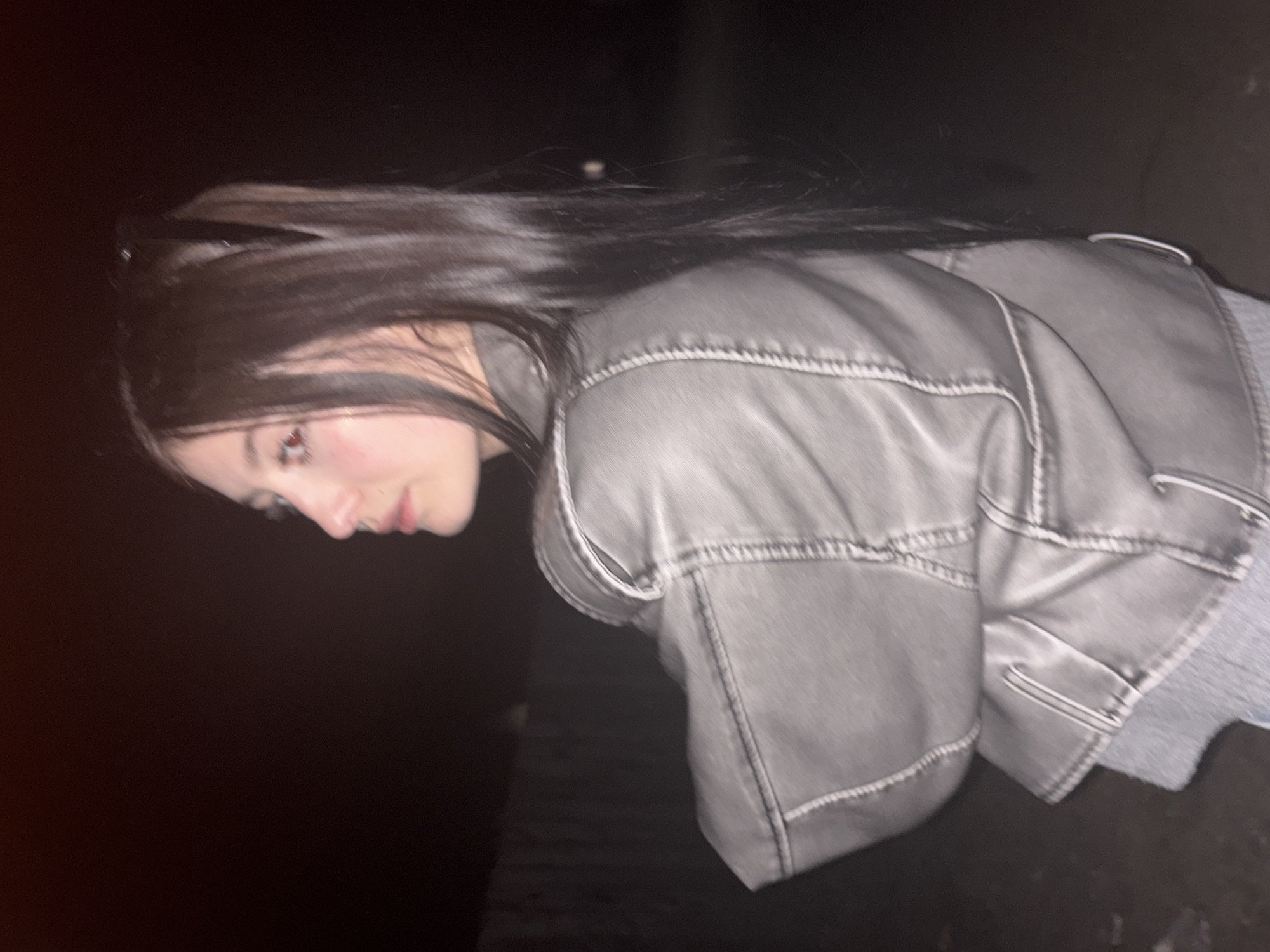 The image shows a person, likely female, with long dark hair in a high ponytail, wearing a grey jacket and a grey top underneath. The person is looking downwards and to the left. The background is dark, suggesting a nighttime setting. There is no indication of location, Russia, or the city of Фоки.