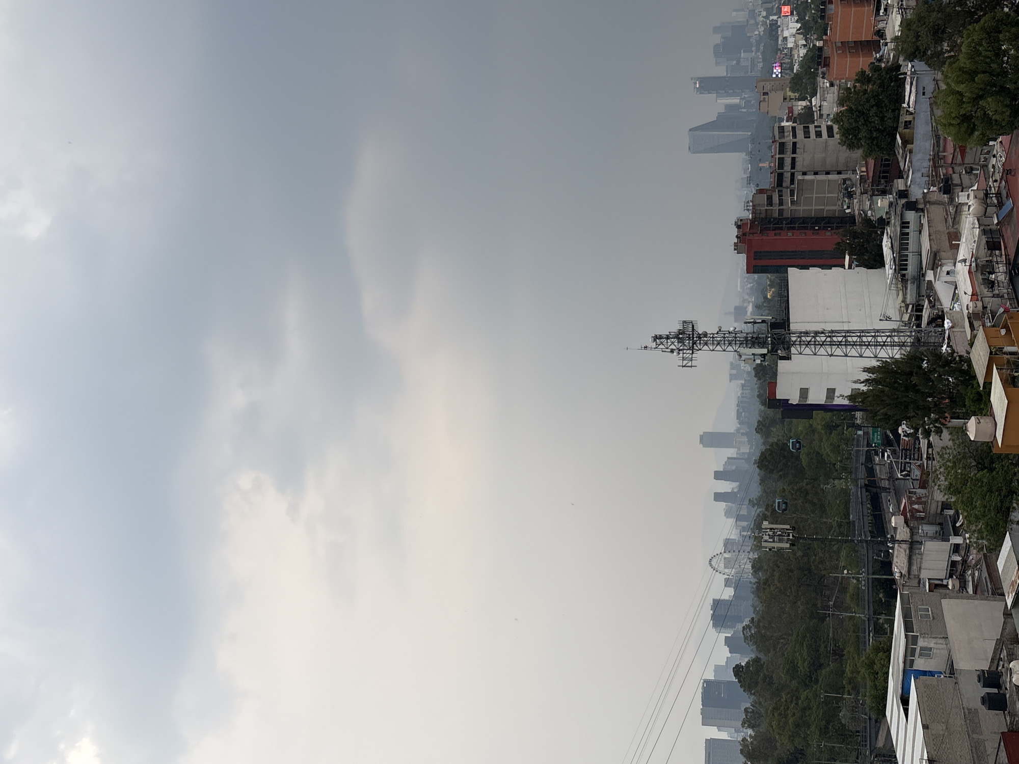 A photograph of the Mexico City skyline during a gray overcast day. A large telecommunications tower sits in the middle ground of the image, with the skyline beyond. A cable car line runs through the middle of the scene with cars suspended. The Bosque de Chapultepec lies beyond, with skyscrapers in the distance and buildings in the foreground.