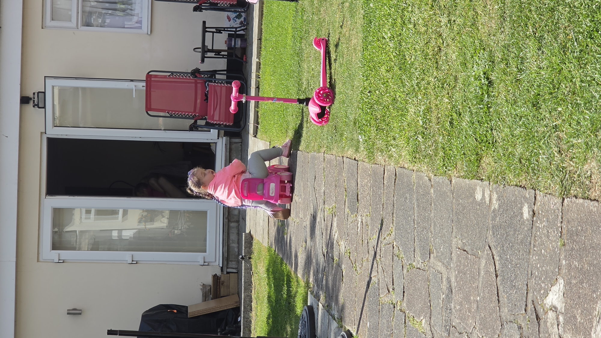 A young girl with braided hair and a pink jacket sits on a pink ride-on toy on a paved area adjacent to a grassy lawn. To the right of the frame, a pink scooter lies on the grass. The paved area appears to be a pathway along the side of a house with white-framed glass doors. In the background, there are items such as folded red chairs and a window. It is daytime with strong sunlight casting shadows on the paving stones. There is no indication of the scene's location in London, United Kingdom.