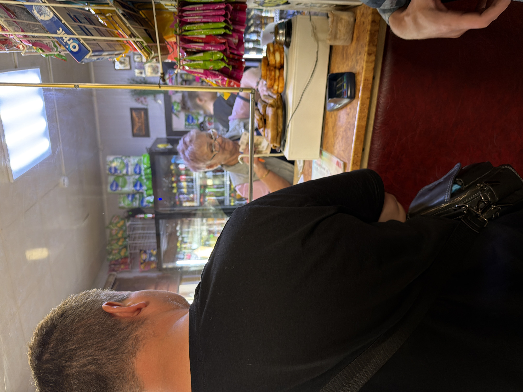 The image appears to be taken inside a small store. In the foreground, a person with short brown hair wearing a black shirt is visible. A black purse is on the right. Through a plastic barrier, an older woman with glasses is seen behind the counter. The store sells various items, including snacks and drinks, which are visible on the shelves. The location is potentially in Russia. The woman is possibly working as a cashier.