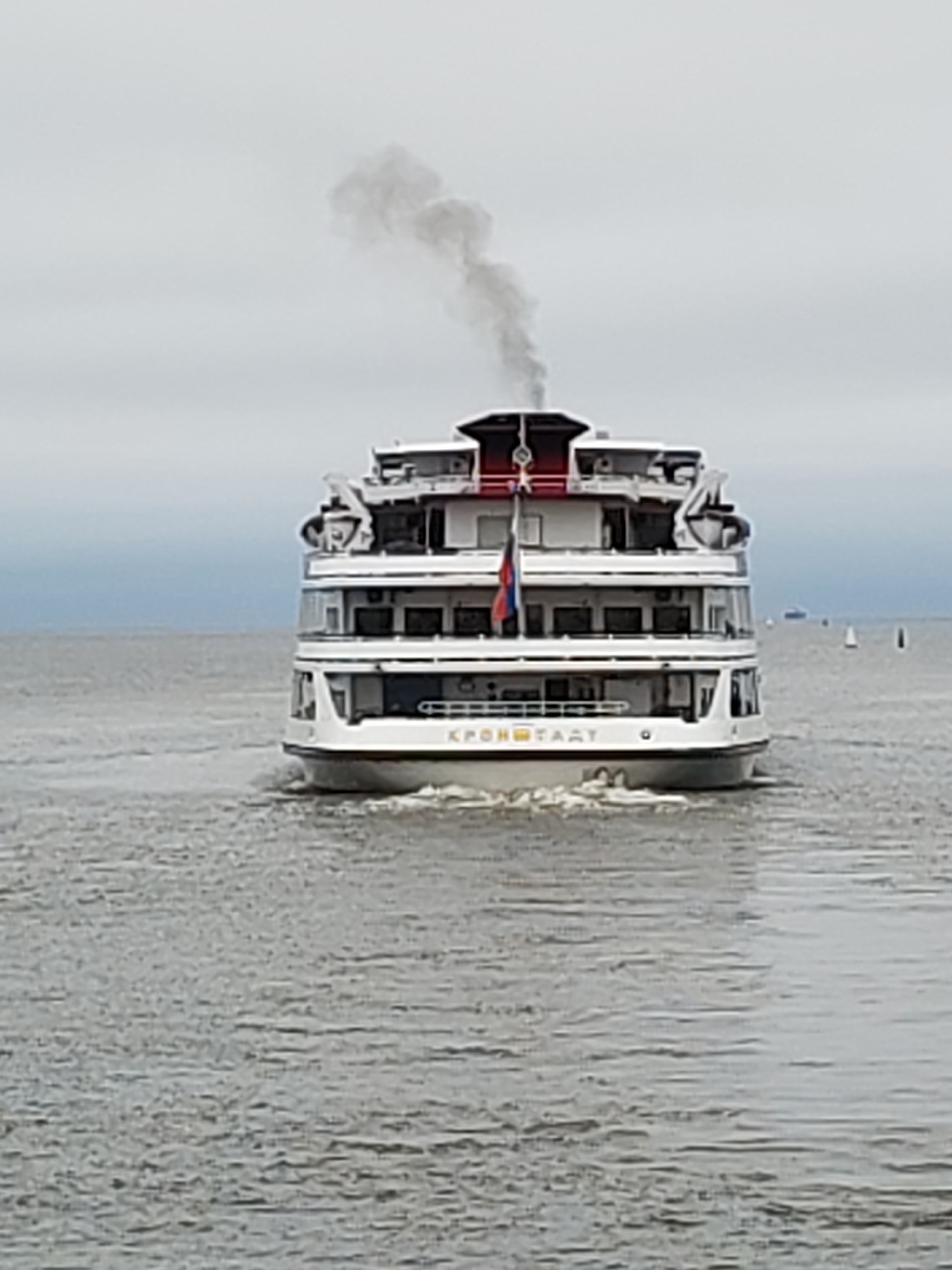 The image shows a white, multi-deck passenger ship sailing on a body of water. The ship has several windows and a red accent on its upper structures. Smoke billows from a smokestack on the top deck. A Russian flag is visible on the side of the ship. The ship is labeled with the Cyrillic text "КРОНШТАДТ" (Kronstadt) on its hull. The water appears choppy, and the sky is overcast. In the distance, other smaller vessels and buoys can be seen. The scene depicts the ship moving through the water in what is likely an industrial or port area, given the presence of a large ship and the overcast weather conditions often associated with such environments.